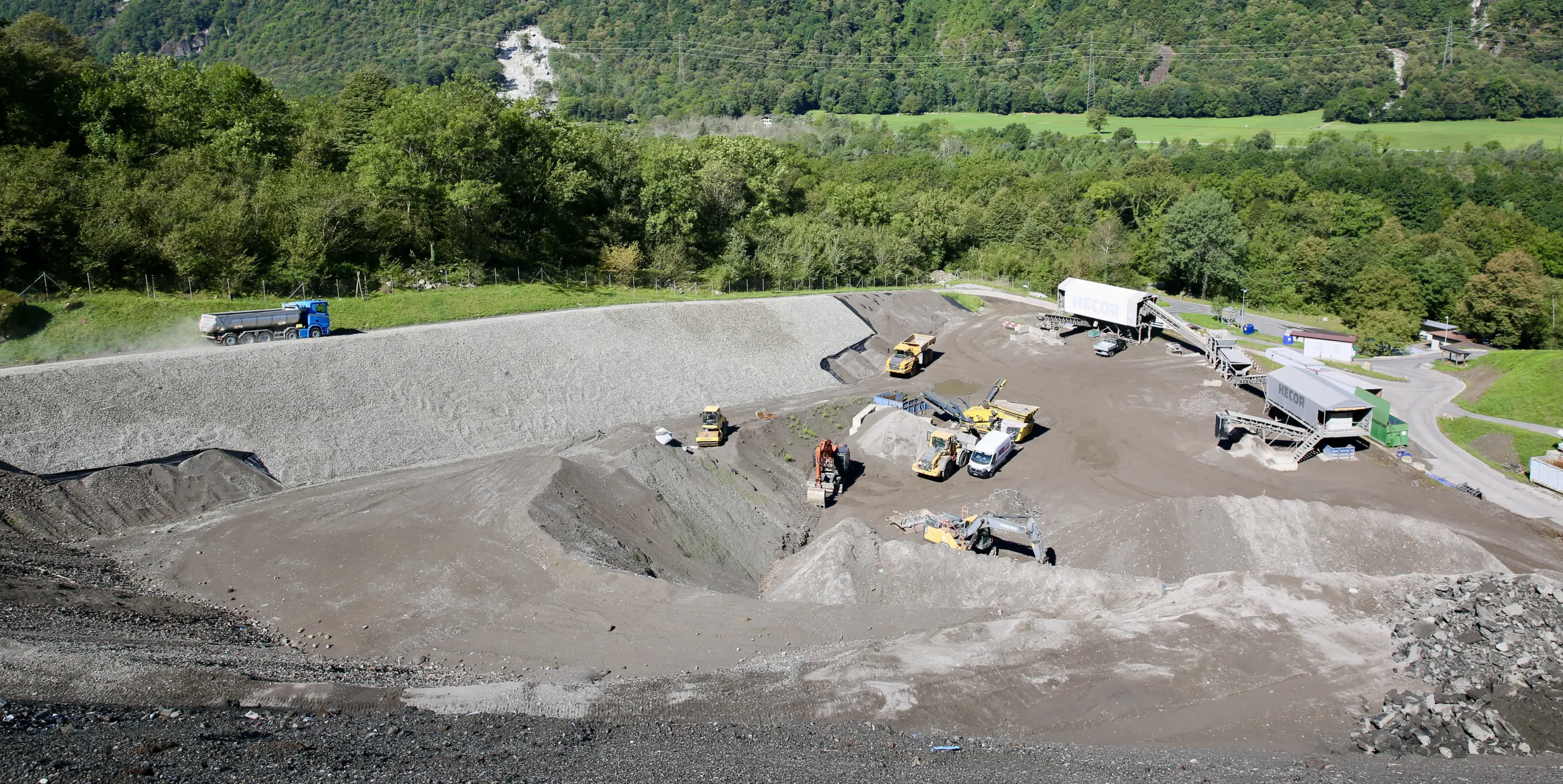 Un cantiere di costruzione con vari macchinari pesanti in azione su un terreno ghiaioso. Si vedono camion, escavatori e una vasta area di terreno lavorato circondata da alberi verdi e colline.