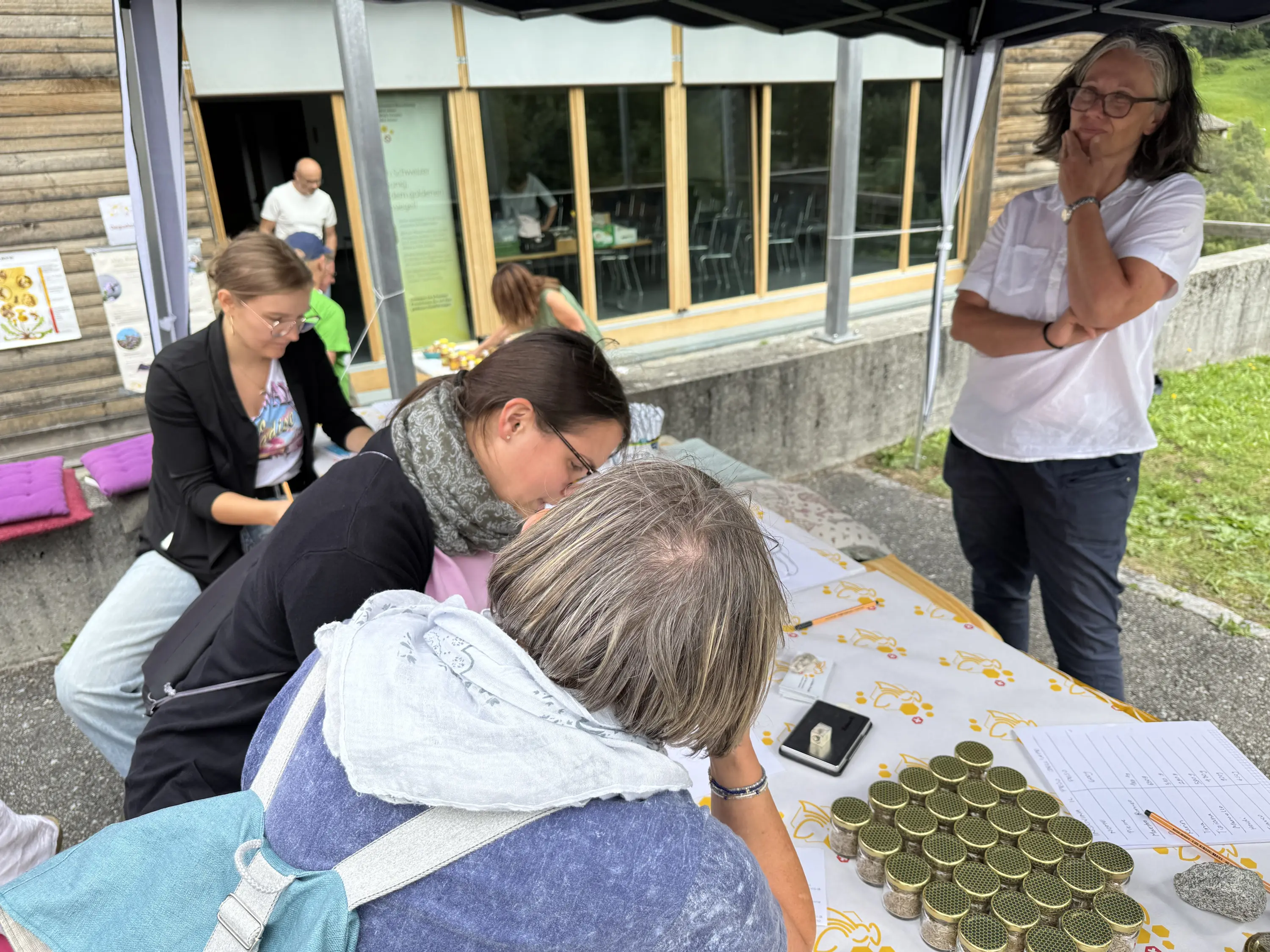 Persone che scrivono e interagiscono attorno a un tavolo all'aperto, con barattoli e materiali disposti davanti a loro. Sullo sfondo, una struttura in legno e persone che partecipano all'evento.