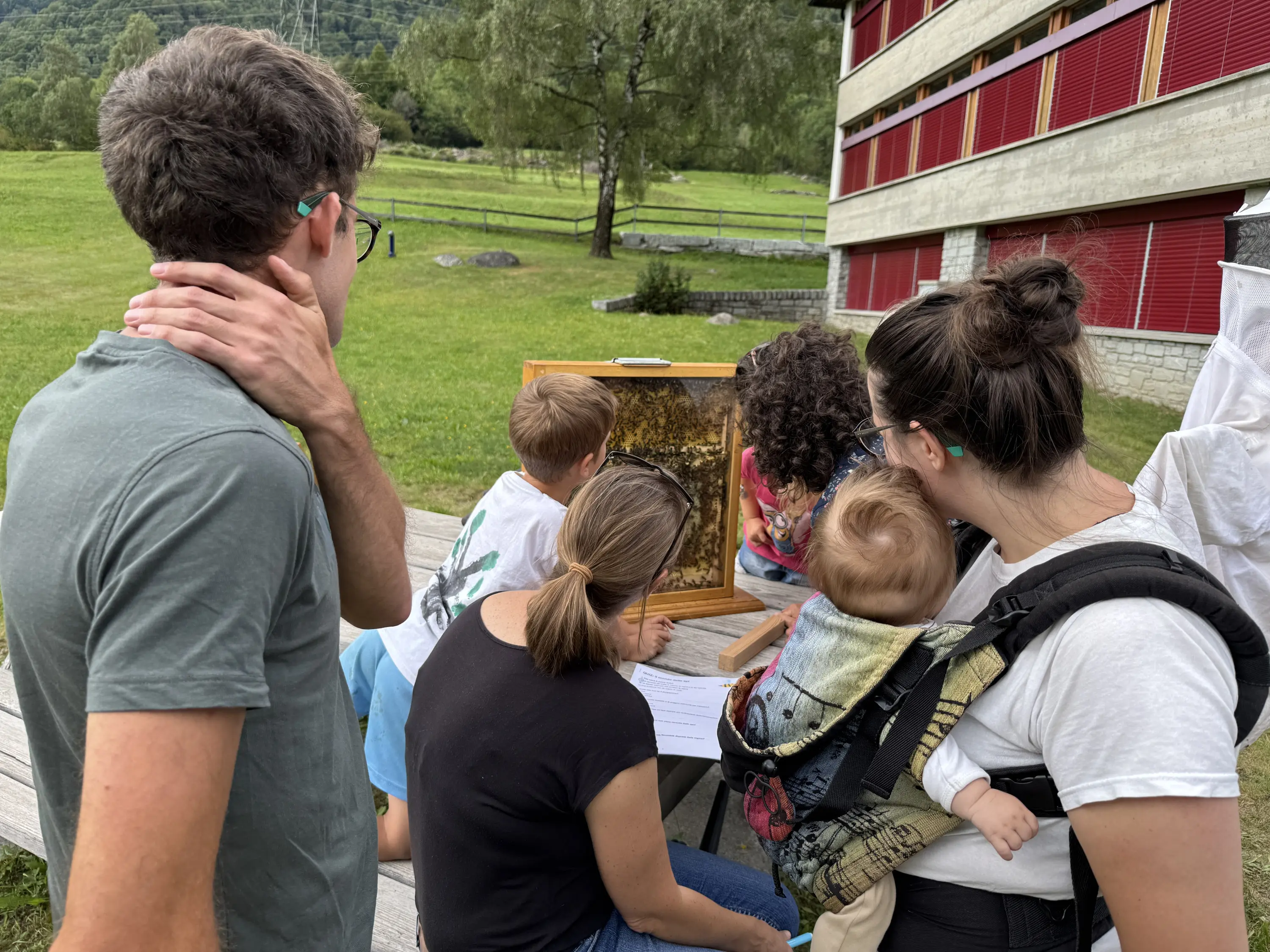 Un gruppo di persone osserva un tavolo con un'immagine, circondato da un paesaggio verde. Alcuni bambini sono presenti, uno di loro è in fasce. I partecipanti sembrano coinvolti e curiosi.