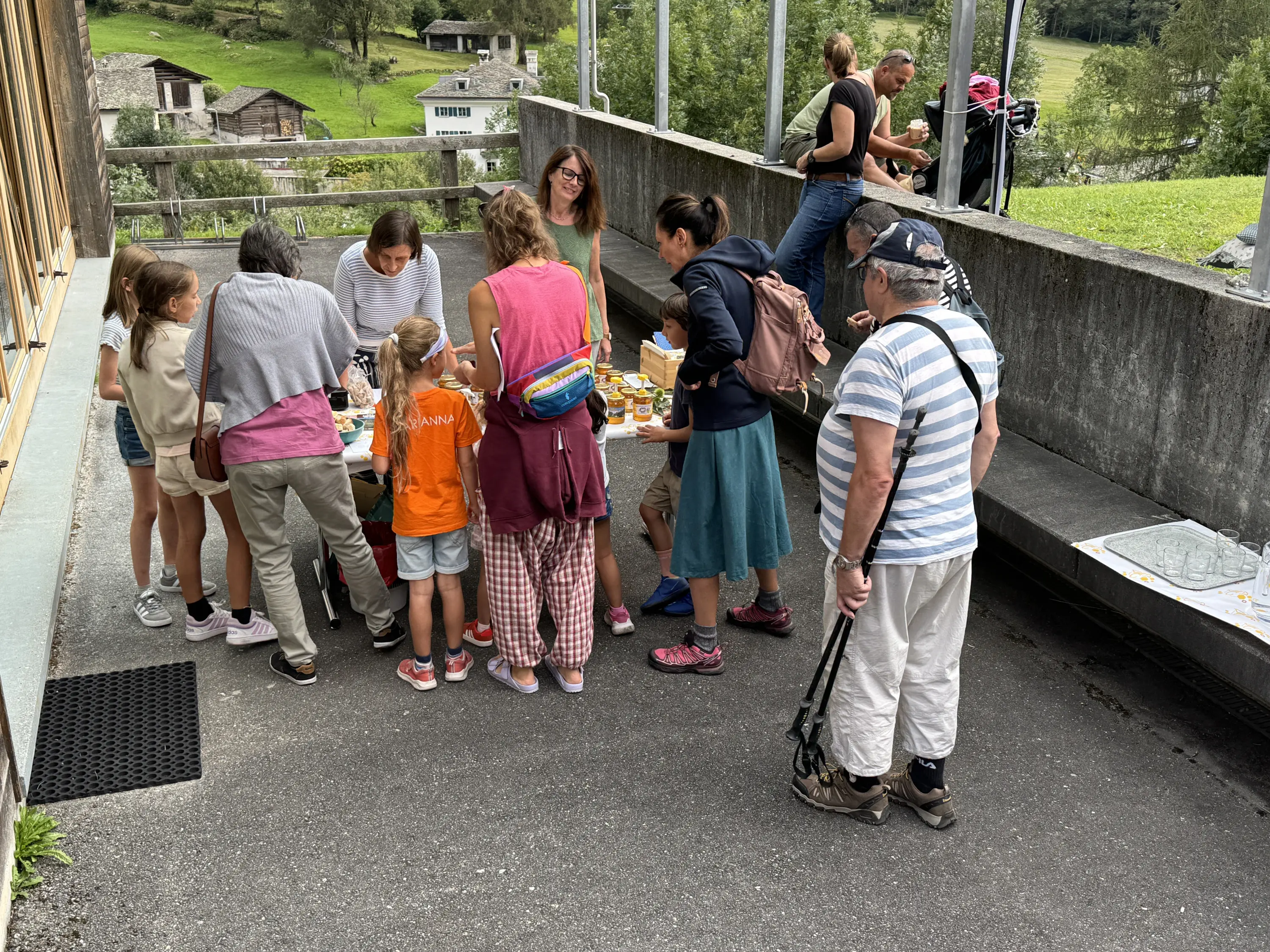 Gruppo di adulti e bambini che si riuniscono attorno a un tavolo all'esterno, con cibo e bevande. Alcuni partecipanti sono seduti e altri in piedi, immersi in conversazione. Sullo sfondo si vedono edifici e natura circostante.