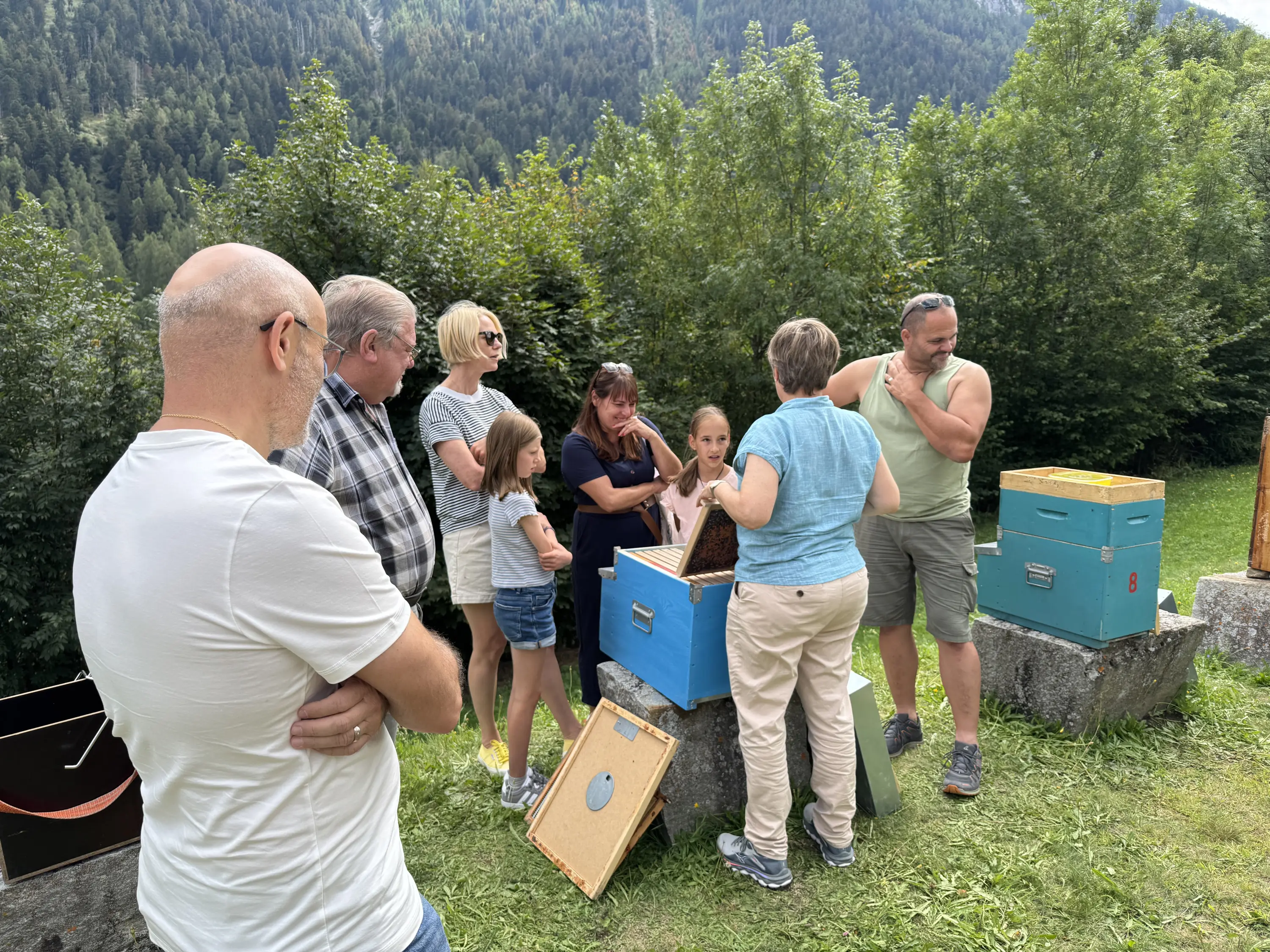 Un gruppo di adulti e bambini osserva un apicoltore che mostra un'arnia in un ambiente naturale montano, circondati da alberi verdi.