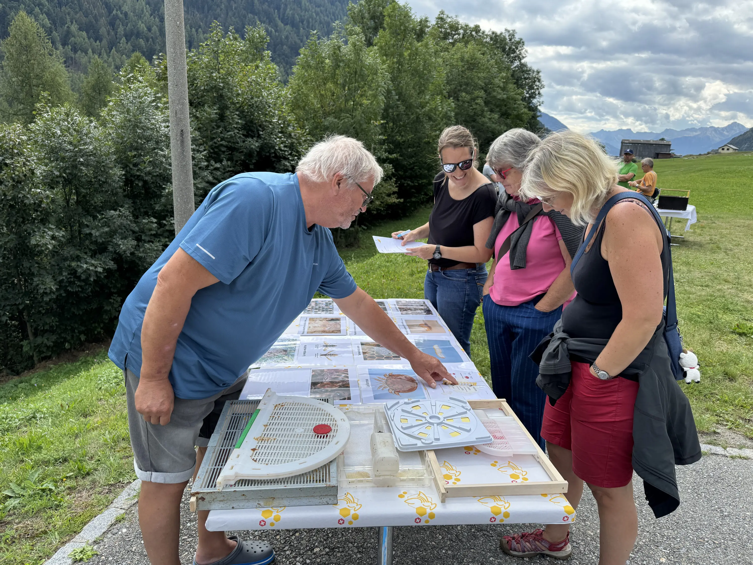 Quattro persone osservano un tavolo con vari materiali, mentre un uomo indica qualcosa. Sullo sfondo si vedono alberi e una collinetta, con un cielo parzialmente nuvoloso.