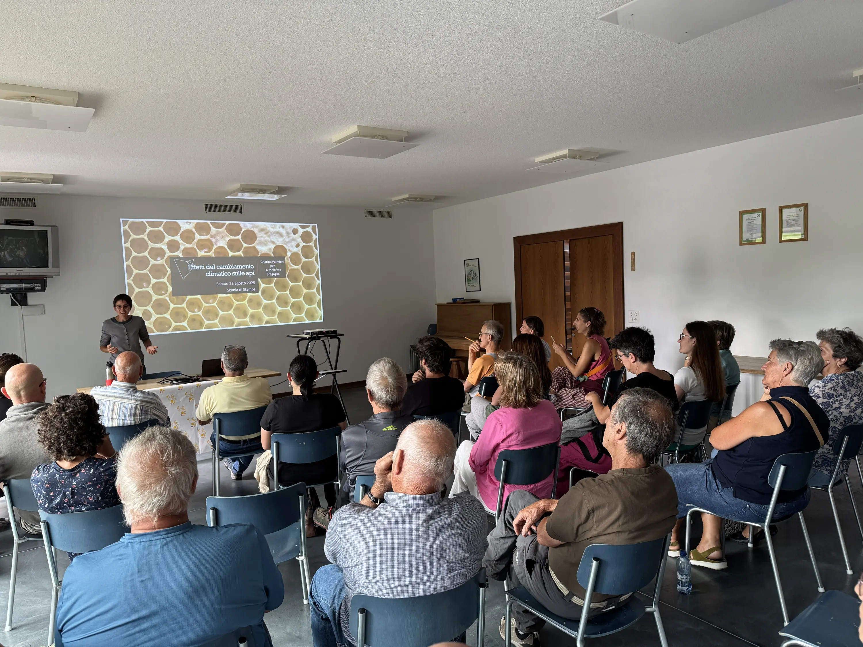 Una sala con diverse persone sedute mentre ascoltano un relatore che presenta un argomento su uno schermo. Il pubblico è composto da adulti di diverse età, alcuni sono attentamente concentrati sul relatore.