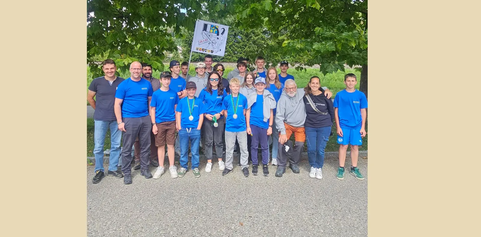 Gruppo di giovani e adulti in magliette blu, sorridenti, posano insieme all'aperto sotto un albero. Alcuni tengono bandiere e medaglie.