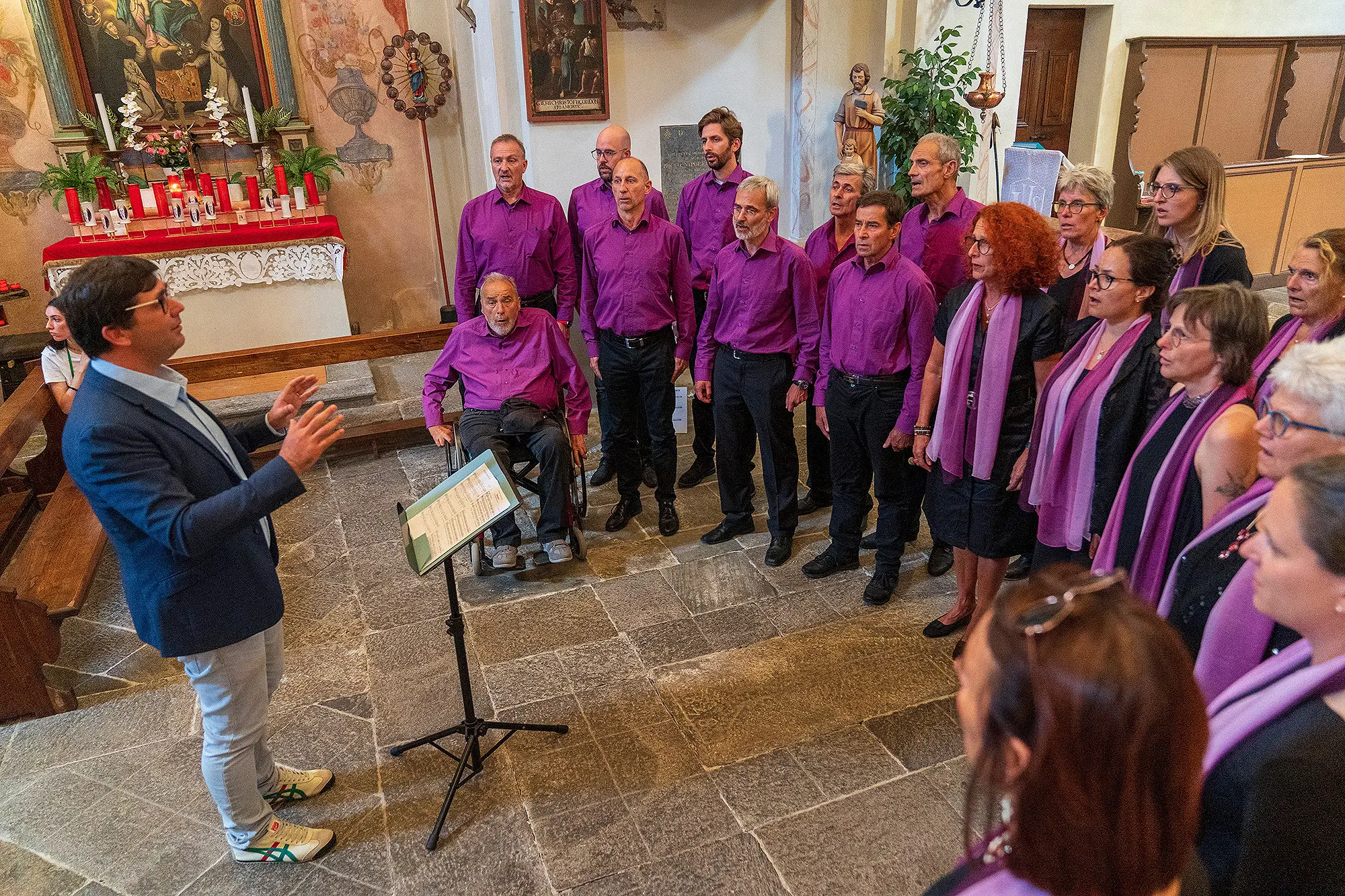 Un coro composto da uomini e donne in abiti viola e sciarpe, si esibisce in una chiesa. Un direttore, in giacca blu, guida il coro, mentre un uomo anziano è seduto sulla sinistra. Sullo sfondo, sono visibili decorazioni religiose e candele.