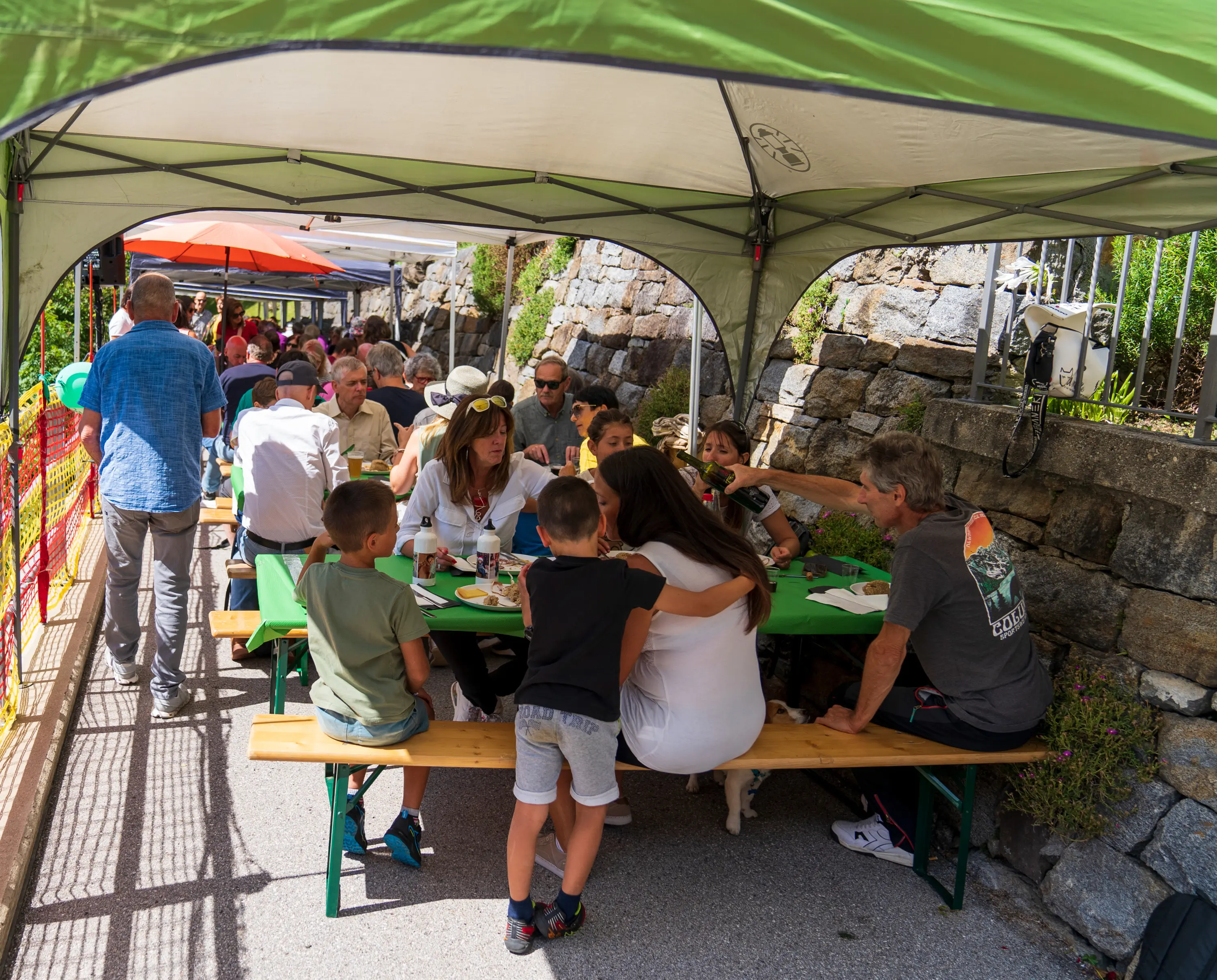 Gruppo di persone sedute a tavoli sotto una tenda, mentre condividono un pasto all'aperto. La scena è vivace, con bambini e adulti che chiacchierano e si divertono insieme.