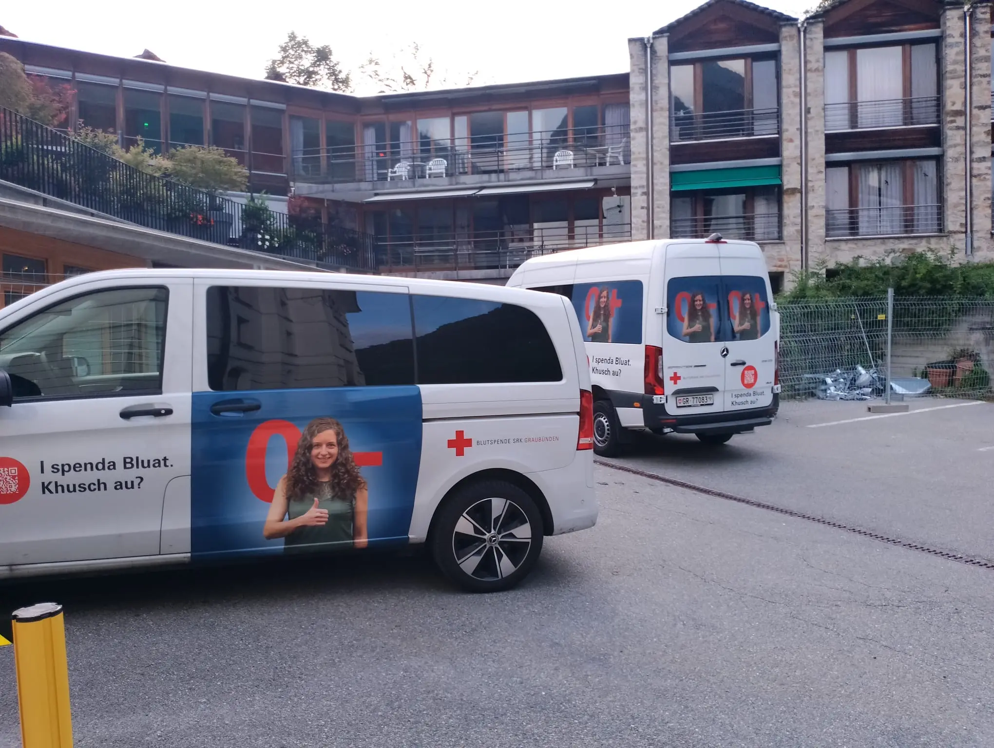 Due furgoni dell'organizzazione di soccorso parcheggiati in un'area esterna di un edificio, con decorazioni visive che promuovono la donazione di sangue.