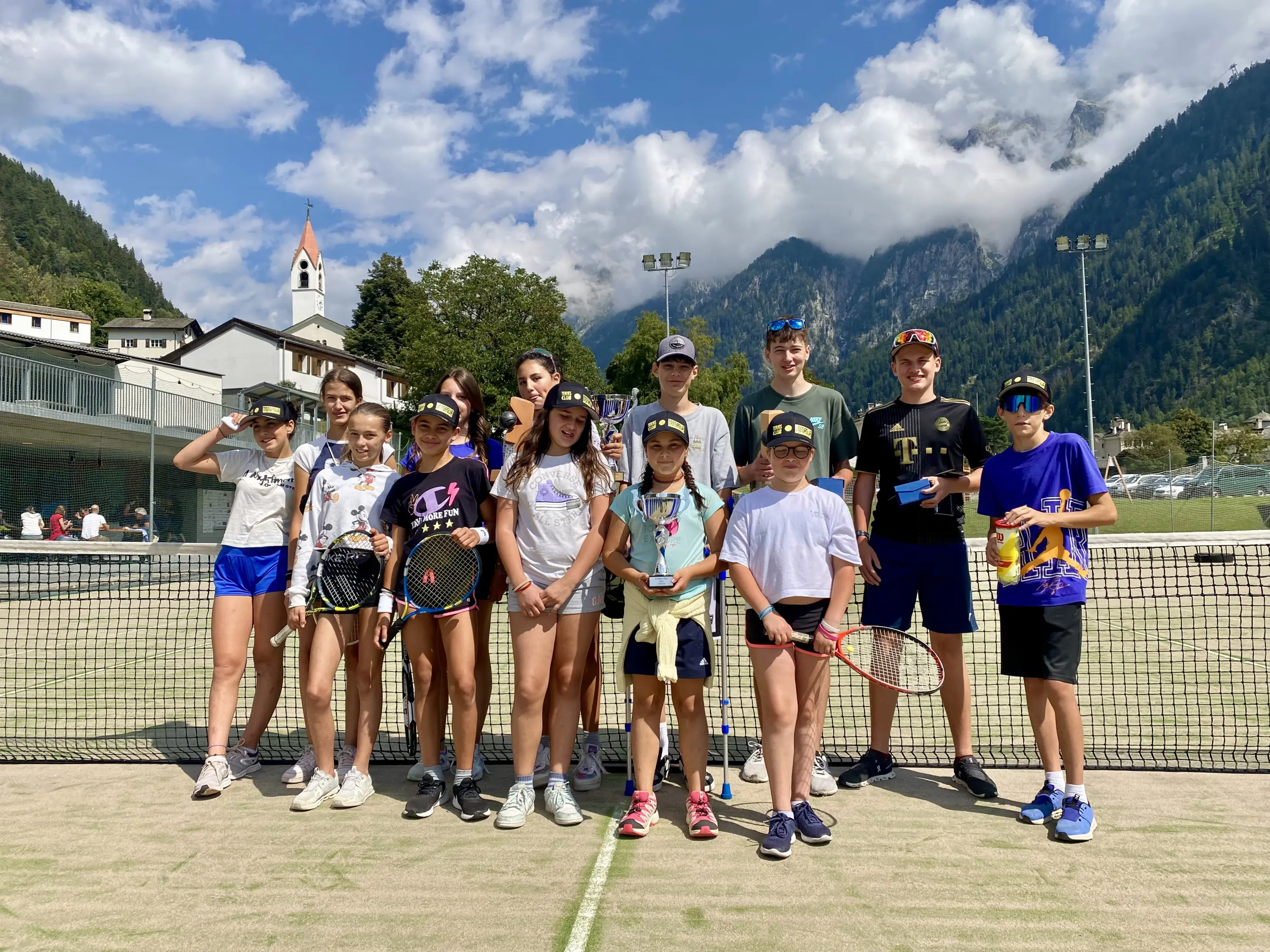 Gruppo di giovani tennisti in campo, sorridenti e pronti per giocare. Sullo sfondo, si vedono montagne e nuvole. Alcuni ragazzi tengono palle e racchette.