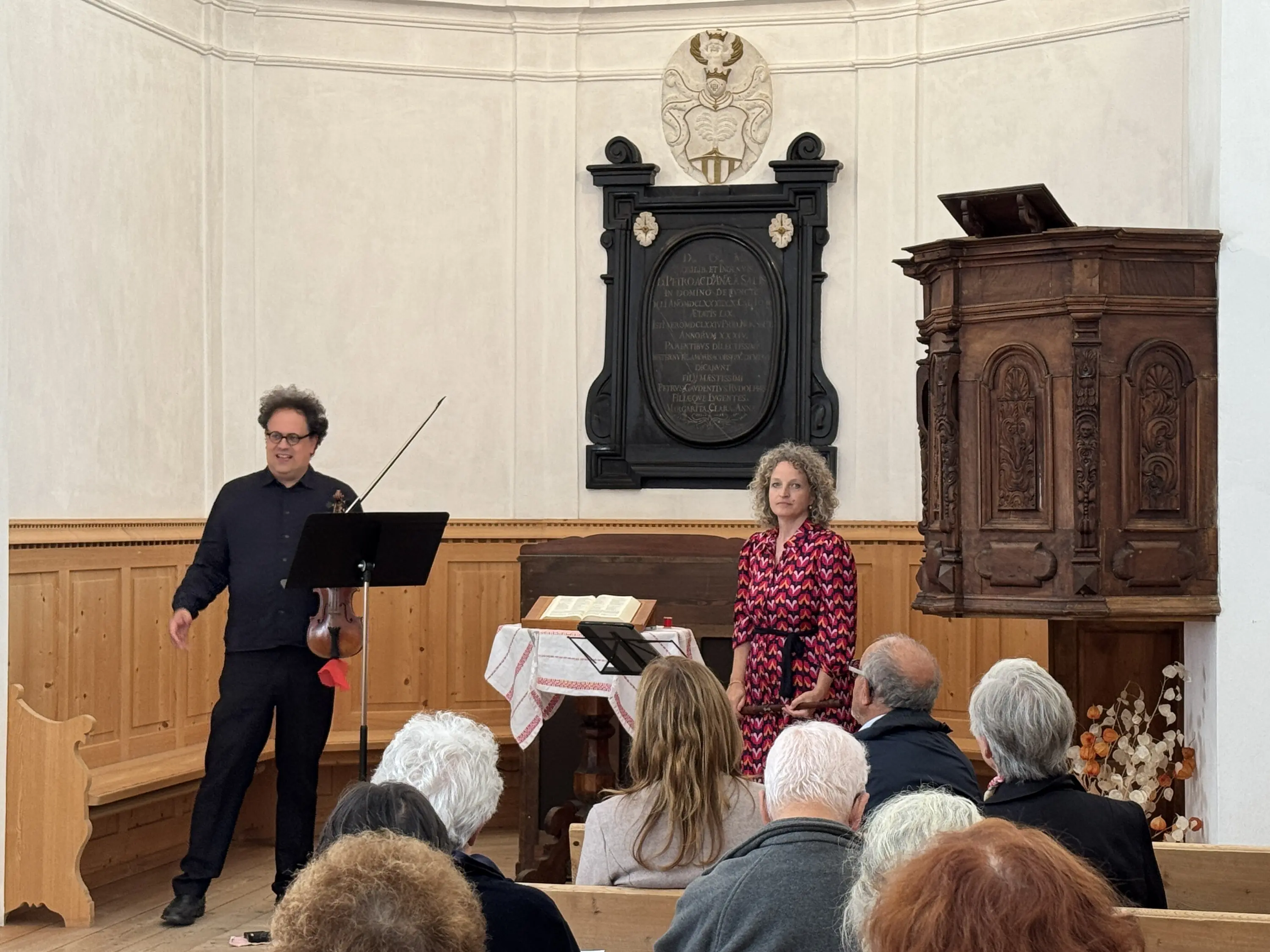 Un violista sta esibendosi in un ambiente ecclesiastico con una donna che parla accanto a lui. Il pubblico è seduto, osservando attentamente. Il luogo presenta arredi in legno e decorazioni storiche.