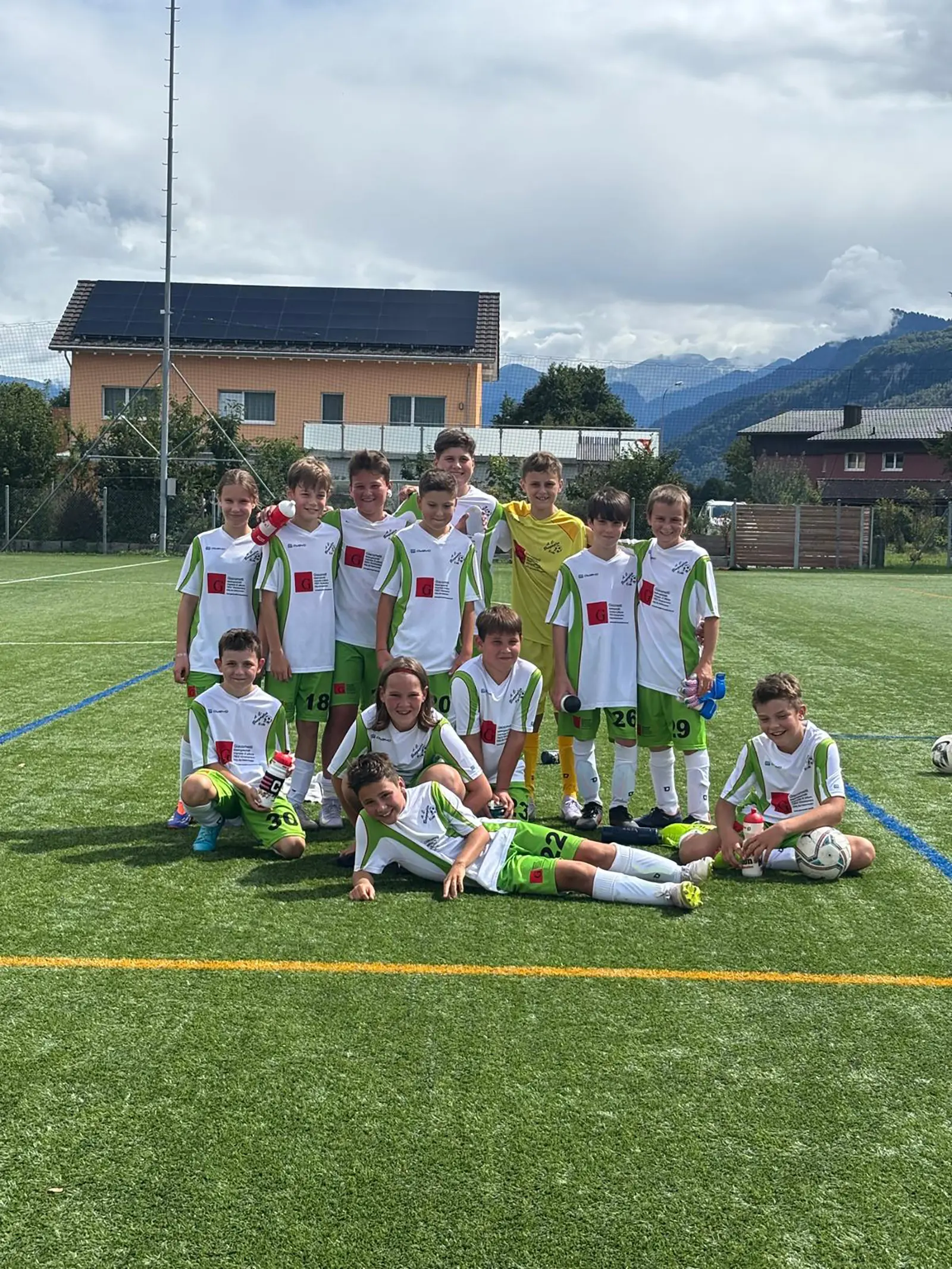 Squadra di calcio giovanile in divisa bianca e verde, posizionata su un campo verde. Alcuni giocatori in piedi, altri accovacciati o sdraiati, sorridono verso la camera. Sullo sfondo si vedono edifici e montagne.