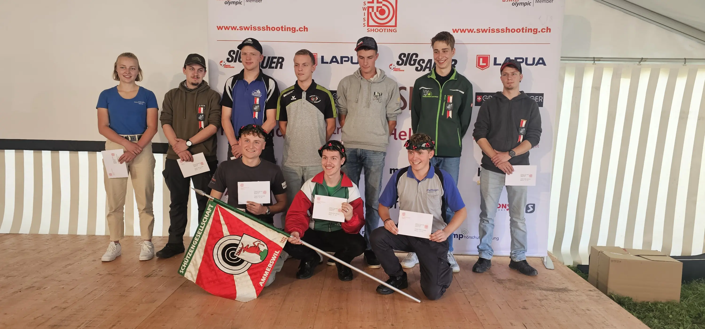 Gruppo di dieci giovani tiratori in un evento sportivo, sorridenti e in posa con trofei e premi. Alcuni indossano magliette di diverse squadre nazionali, inclusa una bandiera. Sullo sfondo, un grande striscione con il logo dell'evento.