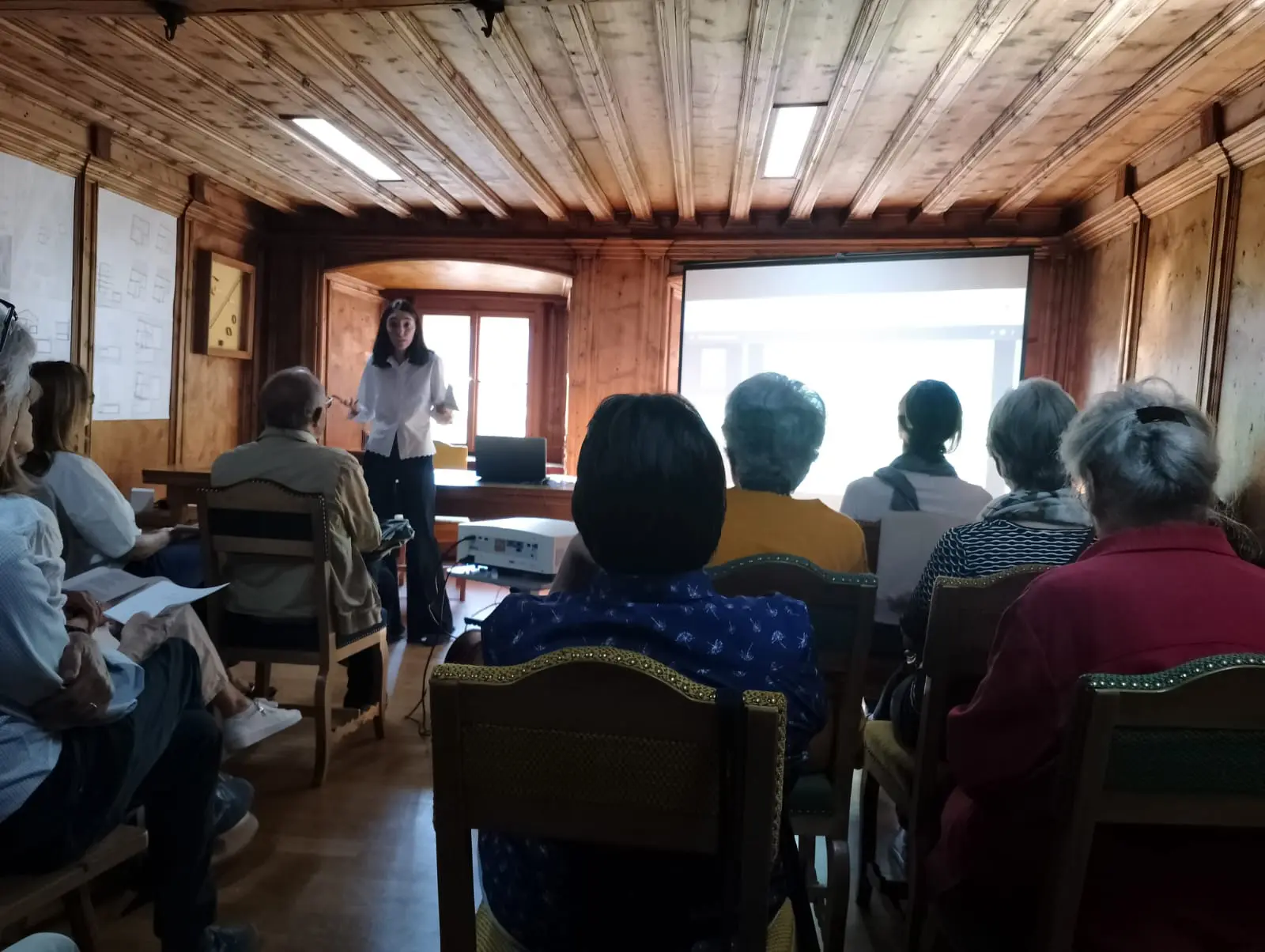 Una presentatrice parla a un gruppo di persone sedute in un'aula con pareti di legno. Un proiettore è visibile sullo sfondo, ma il contenuto proiettato non è leggibile.