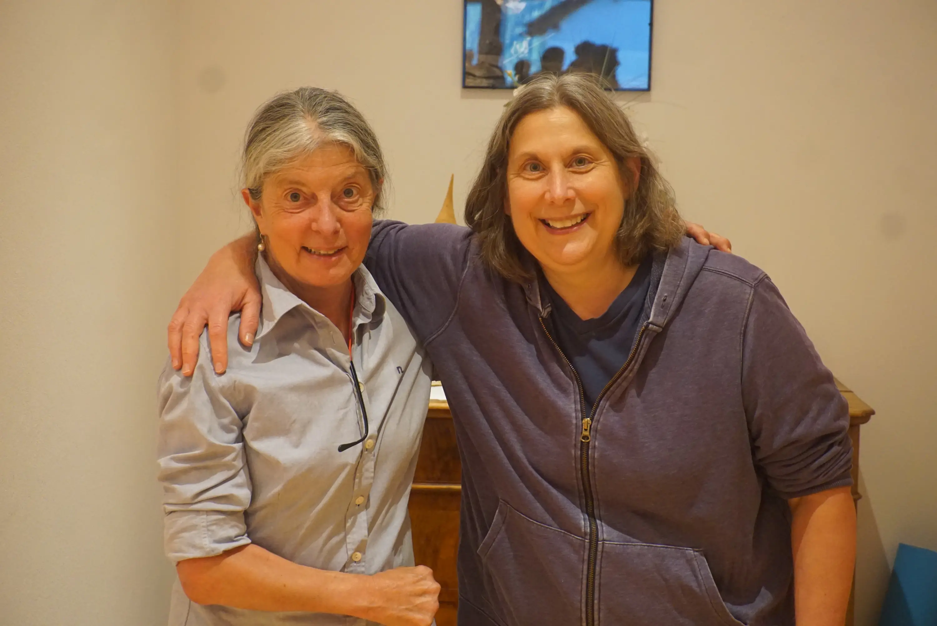 Due donne sorridenti si abbracciano in un ambiente accogliente. Una ha i capelli grigi e indossa una camicia chiara, mentre l'altra ha capelli castani e indossa una felpa viola. Sullo sfondo si intravede un quadro sulla parete.