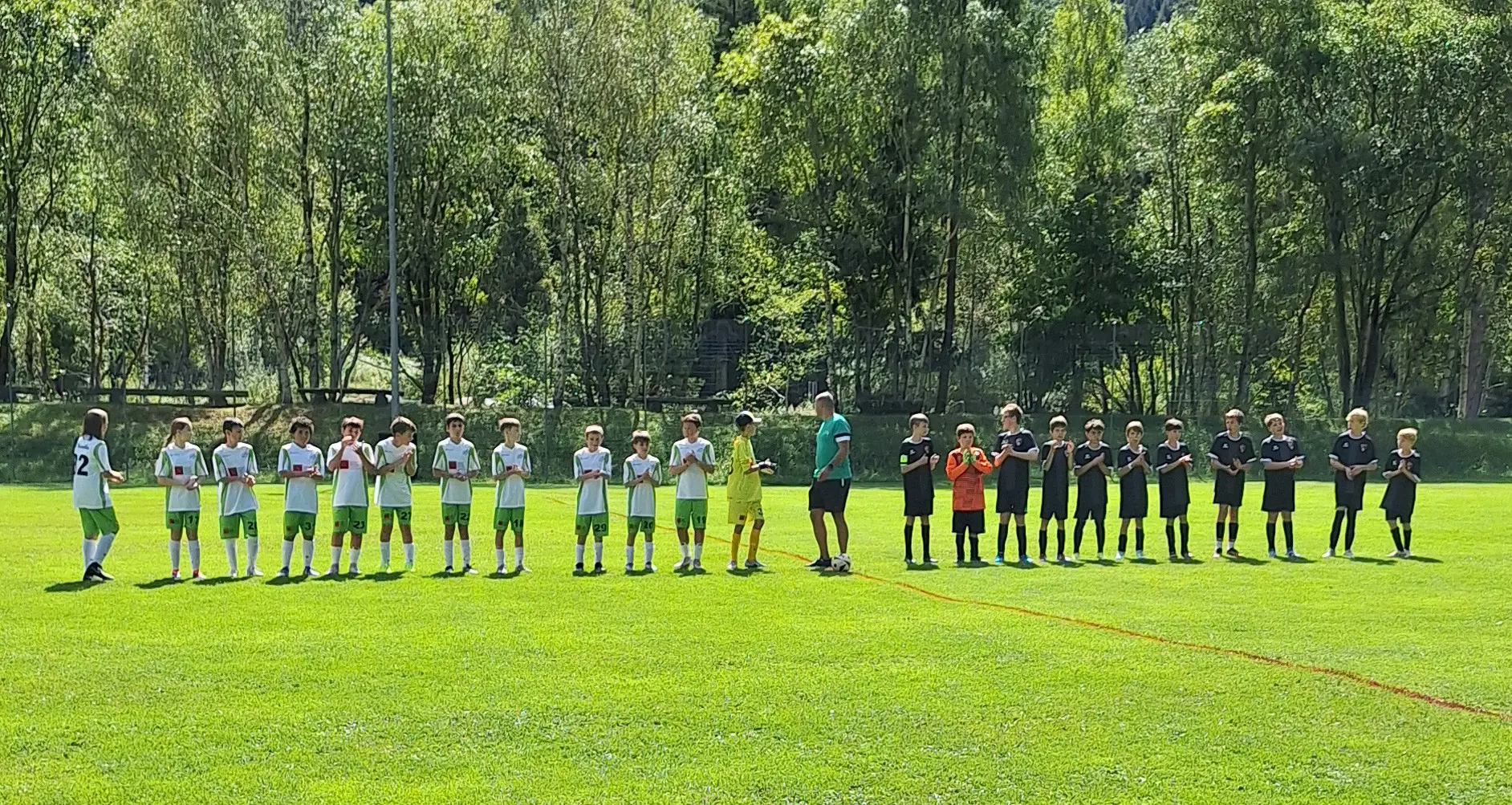 Giocatori di calcio in campo, schierati in due squadre di diverse divise, pronti per una partita. Sullo sfondo, alberi e un campo ben curato.