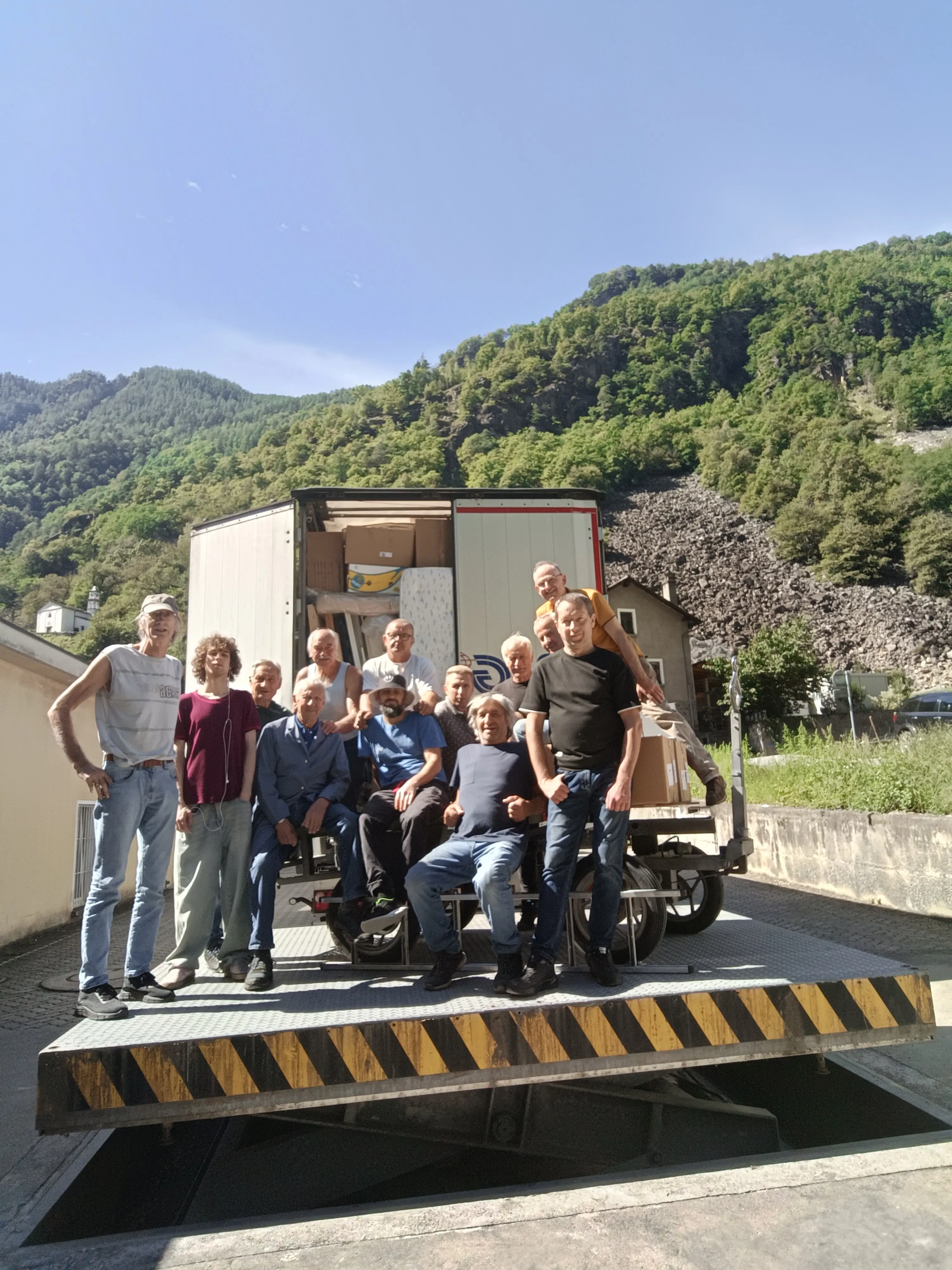 Gruppo di persone in posa su un camion di carico in una zona montana. Sullo sfondo si vedono alberi e rocce. Tutti sorridono e sono vestiti casualmente.