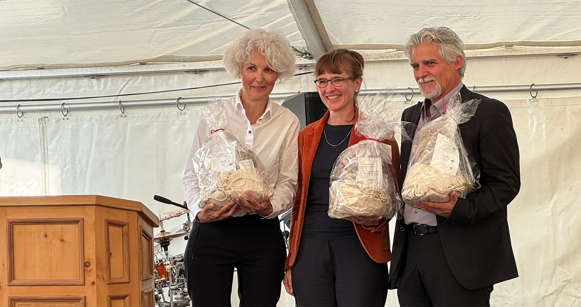 Tre persone posano sorridenti all'interno di una tenda. Ognuno di loro tiene un sacchetto trasparente con all'interno dei premi. Sullo sfondo, si intravedono strumenti musicali e un podio di legno.