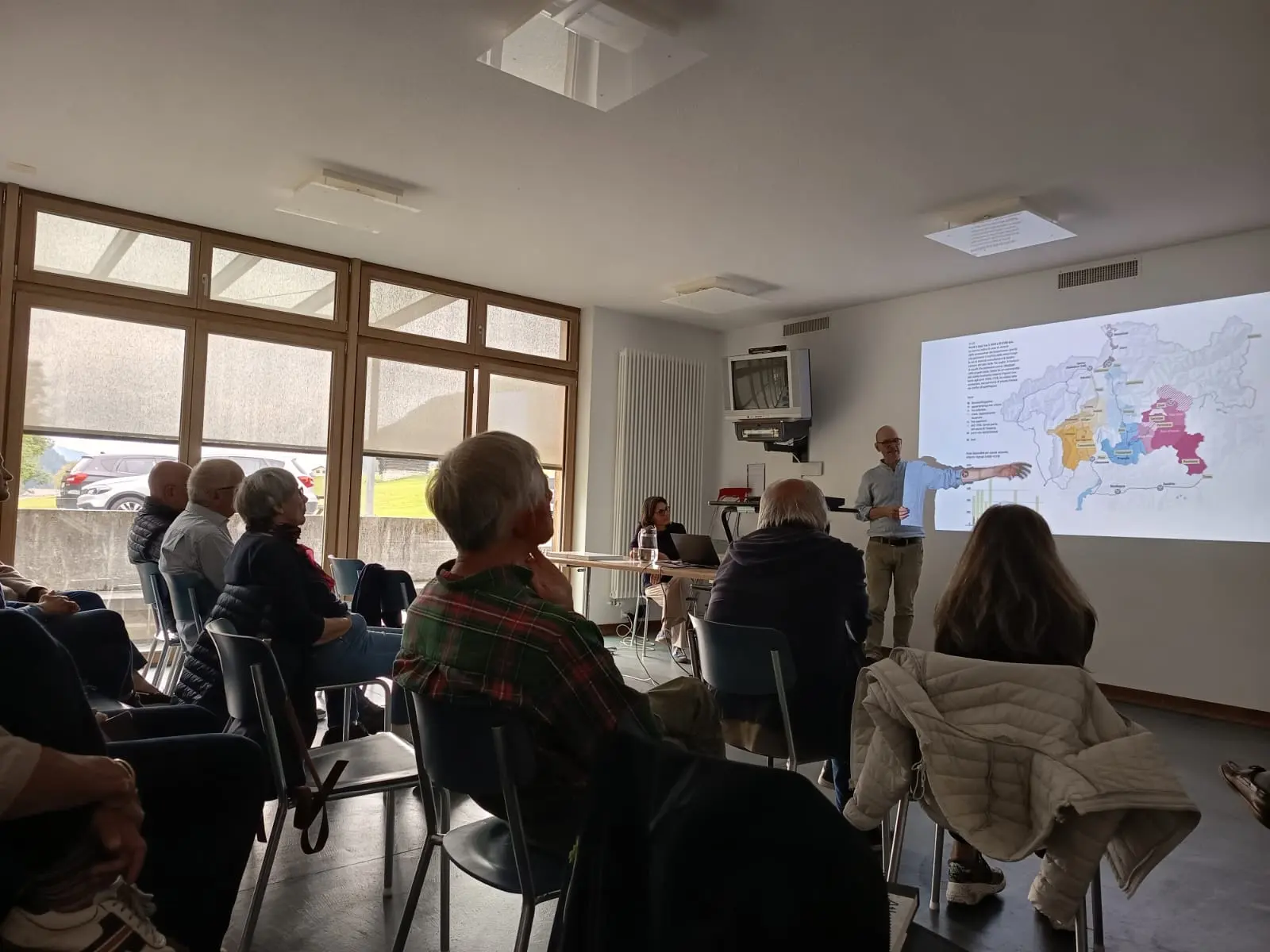 Un gruppo di persone in una sala conferenze ascolta un relatore che presenta una mappa su uno schermo. I partecipanti sono seduti su sedie, mentre alcune finestre lasciano entrare la luce.