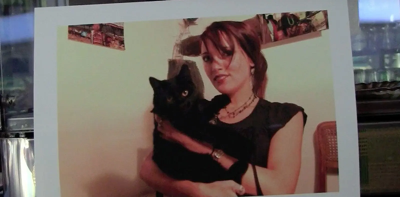 Una giovane donna sorride mentre tiene in braccio un gatto nero. Sullo sfondo ci sono decorazioni murali e una sedia di vimini.