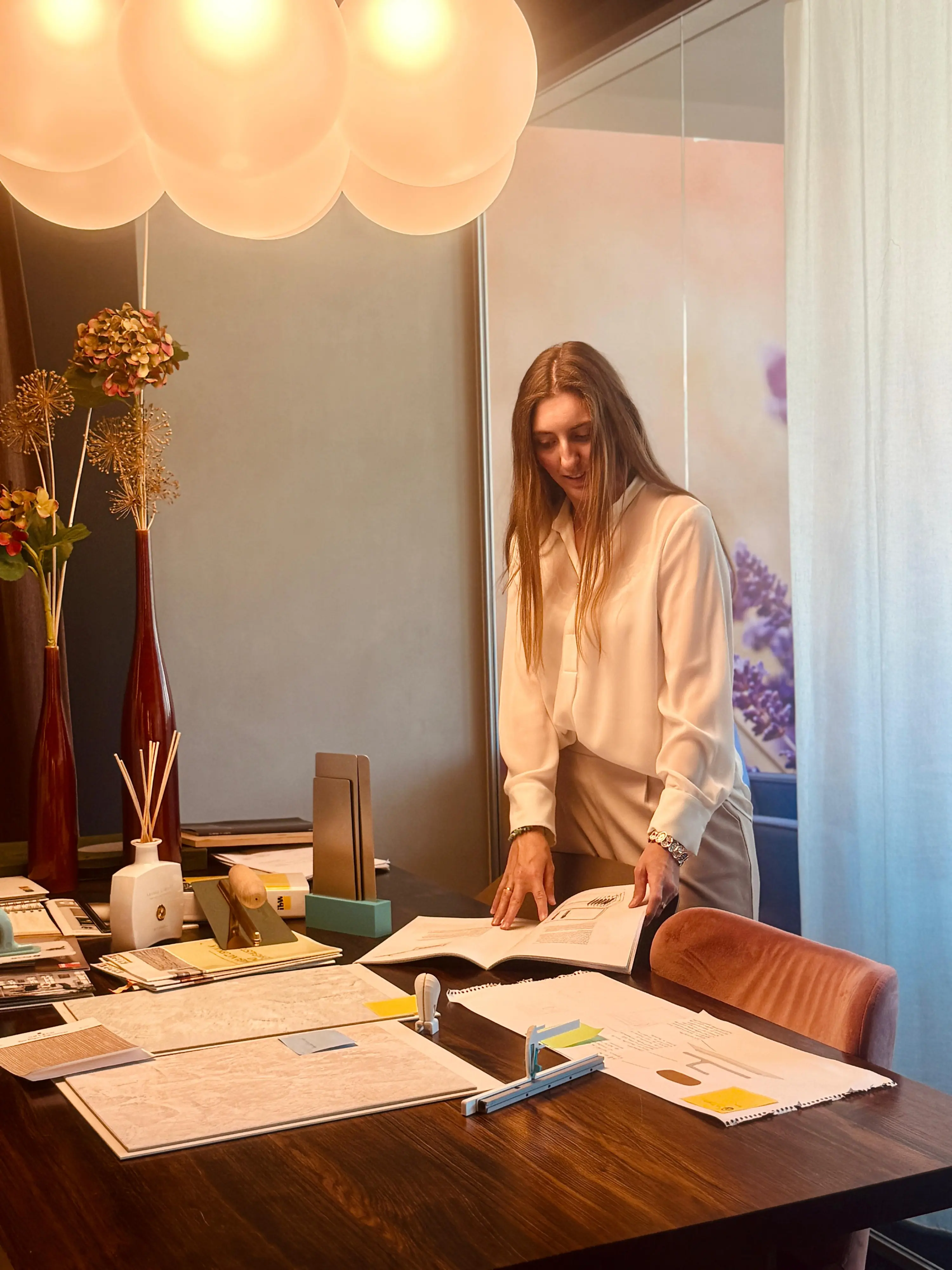 Una donna sorride mentre sfoglia documenti su un tavolo di lavoro elegante, circondata da decorazioni moderne e un'illuminazione calda.