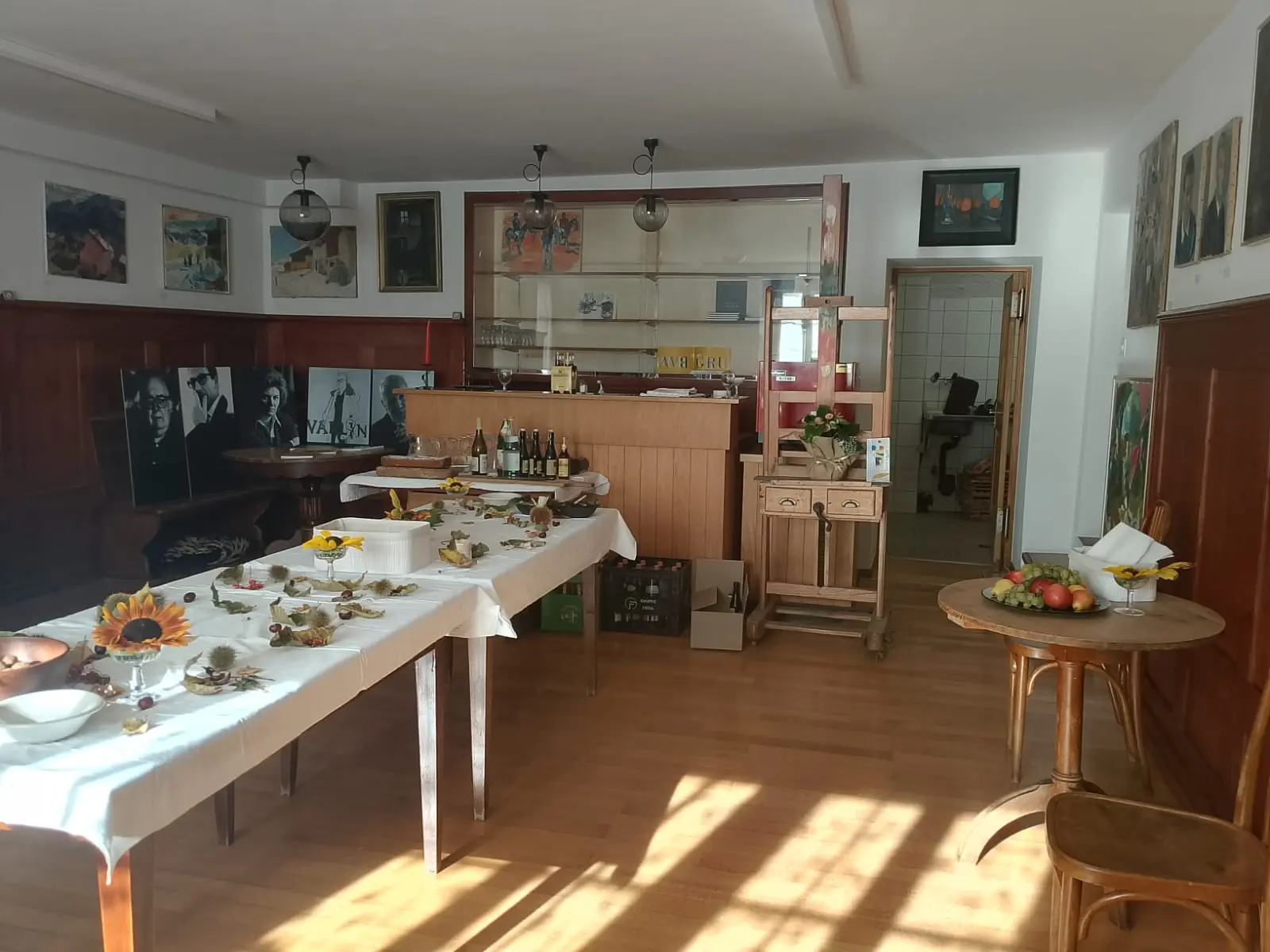 Un ambiente interno con tavoli apparecchiati, cibo e bibite. Sullo sfondo, un bancone e opere d'arte alle pareti. Due tavoli presentano piatti di frutta e decorazioni floreali.