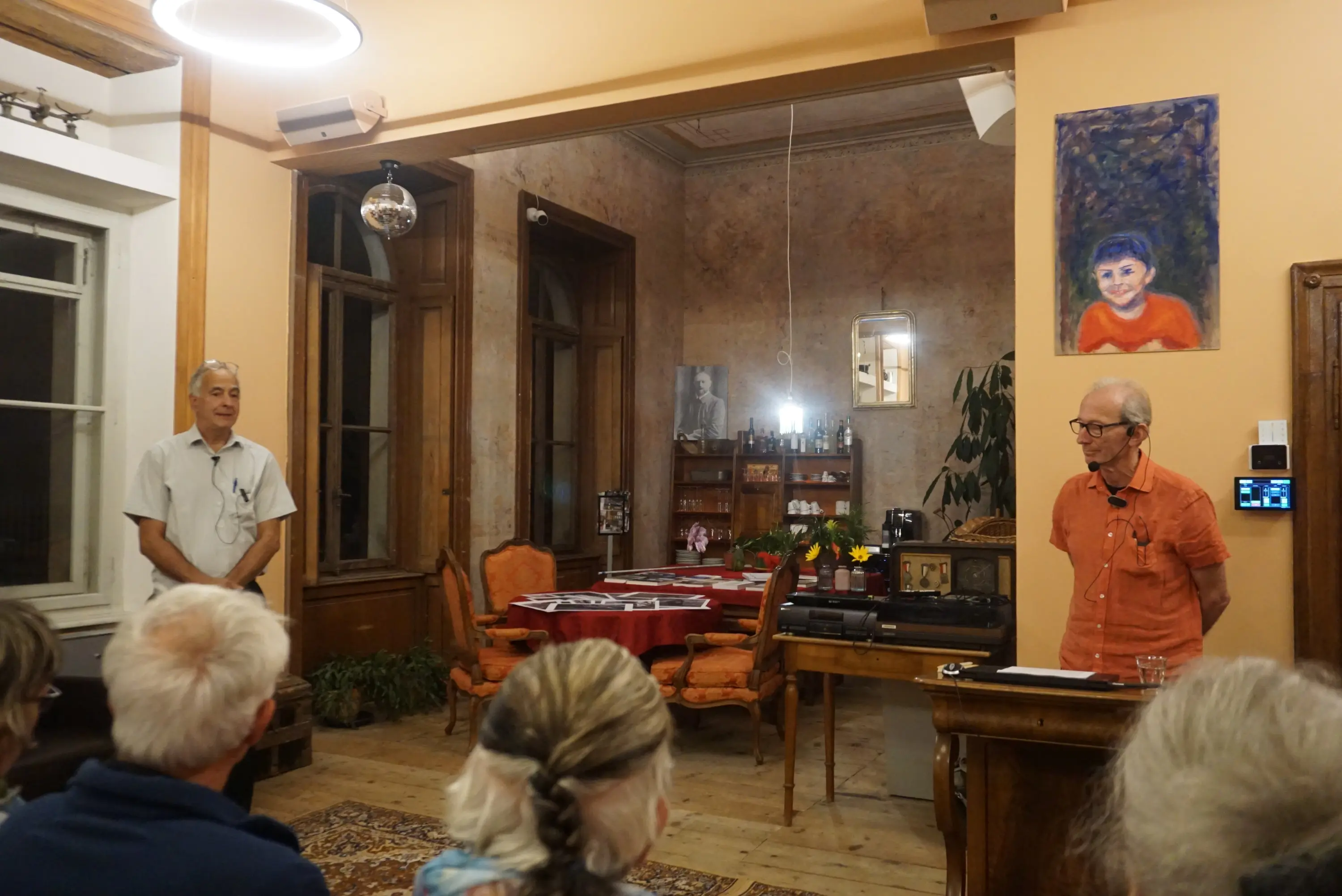 Due persone parlano in una sala accogliente con decorazioni artistiche e un tavolo imbandito sullo sfondo. I partecipanti assistono all'evento seduti in posizione frontale.