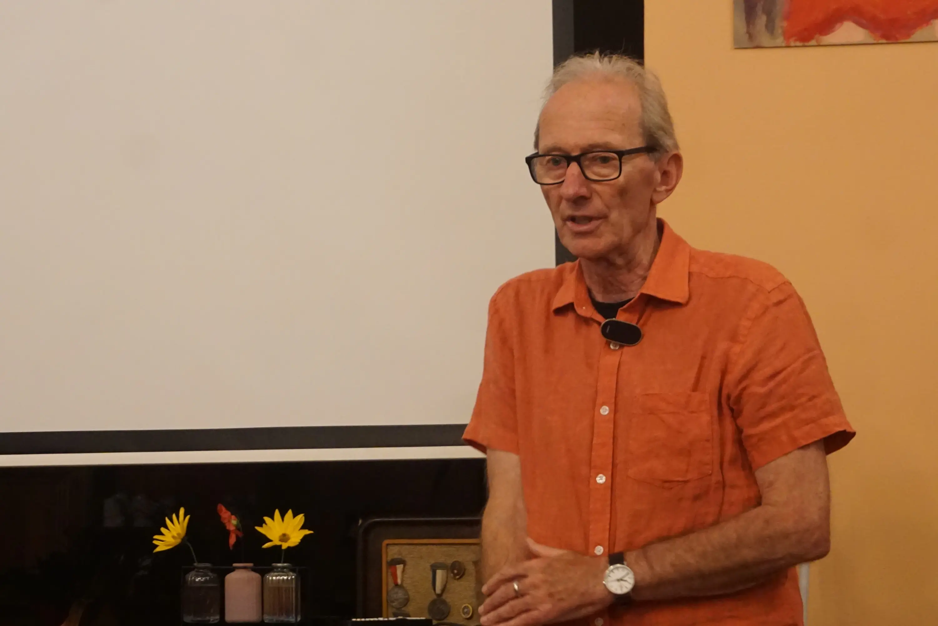 Uomo con occhiali e camicia arancione parla di fronte a uno schermo bianco. Sul tavolo ci sono dei fiori e alcuni oggetti decorativi.