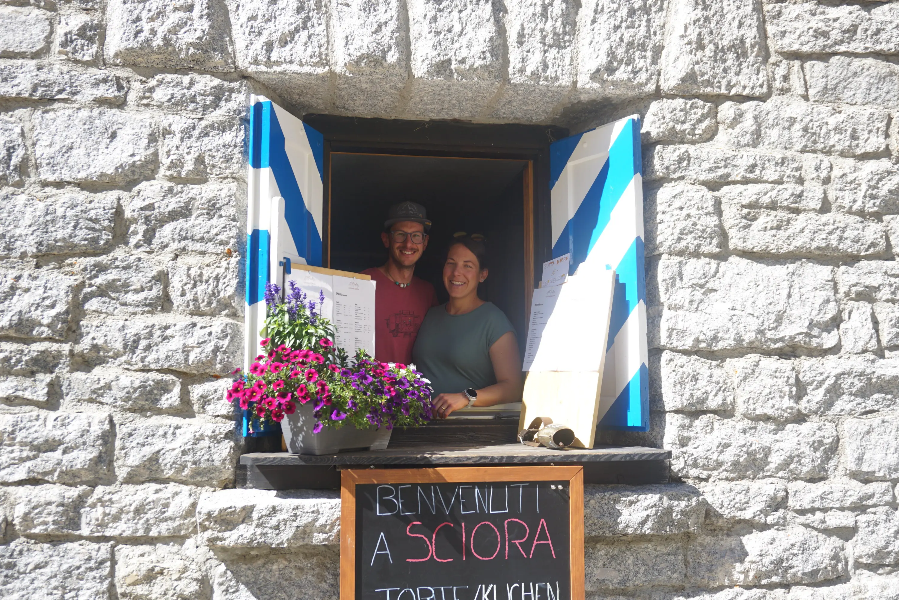 Due persone sorridenti si affacciano da una finestra con decorazioni blu e bianche. Davanti, una lavagna scritta a mano con la scritta "Benvenuti a Sciora". Sono presenti fiori colorati in una fioriera.