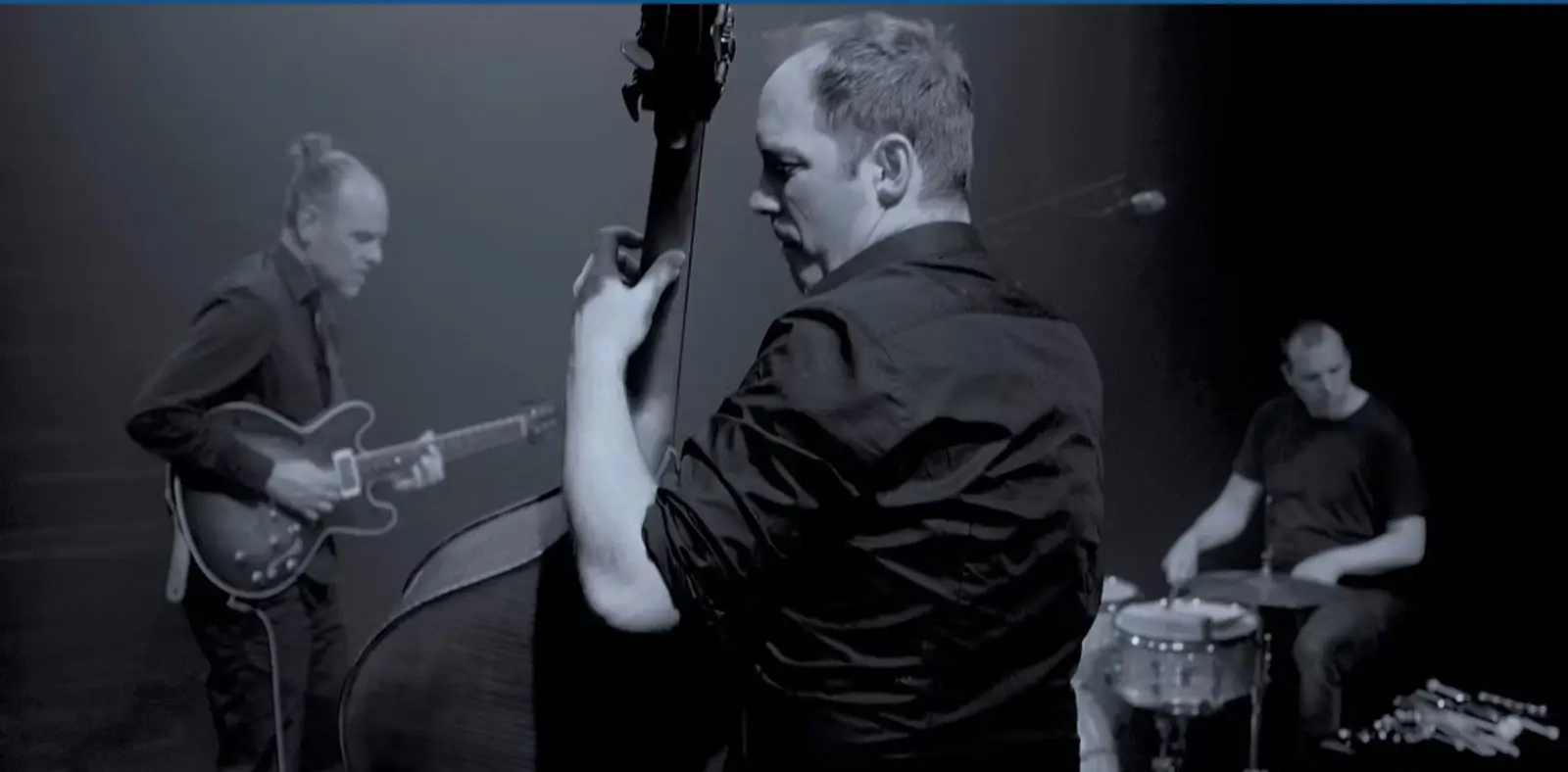 Tre musicisti si esibiscono su un palco illuminato. Un uomo suona la chitarra elettrica, un altro suona il contrabbasso, mentre il terzo è seduto al tamburo. L'atmosfera è intensa e il loro abbigliamento è elegante e scuro.