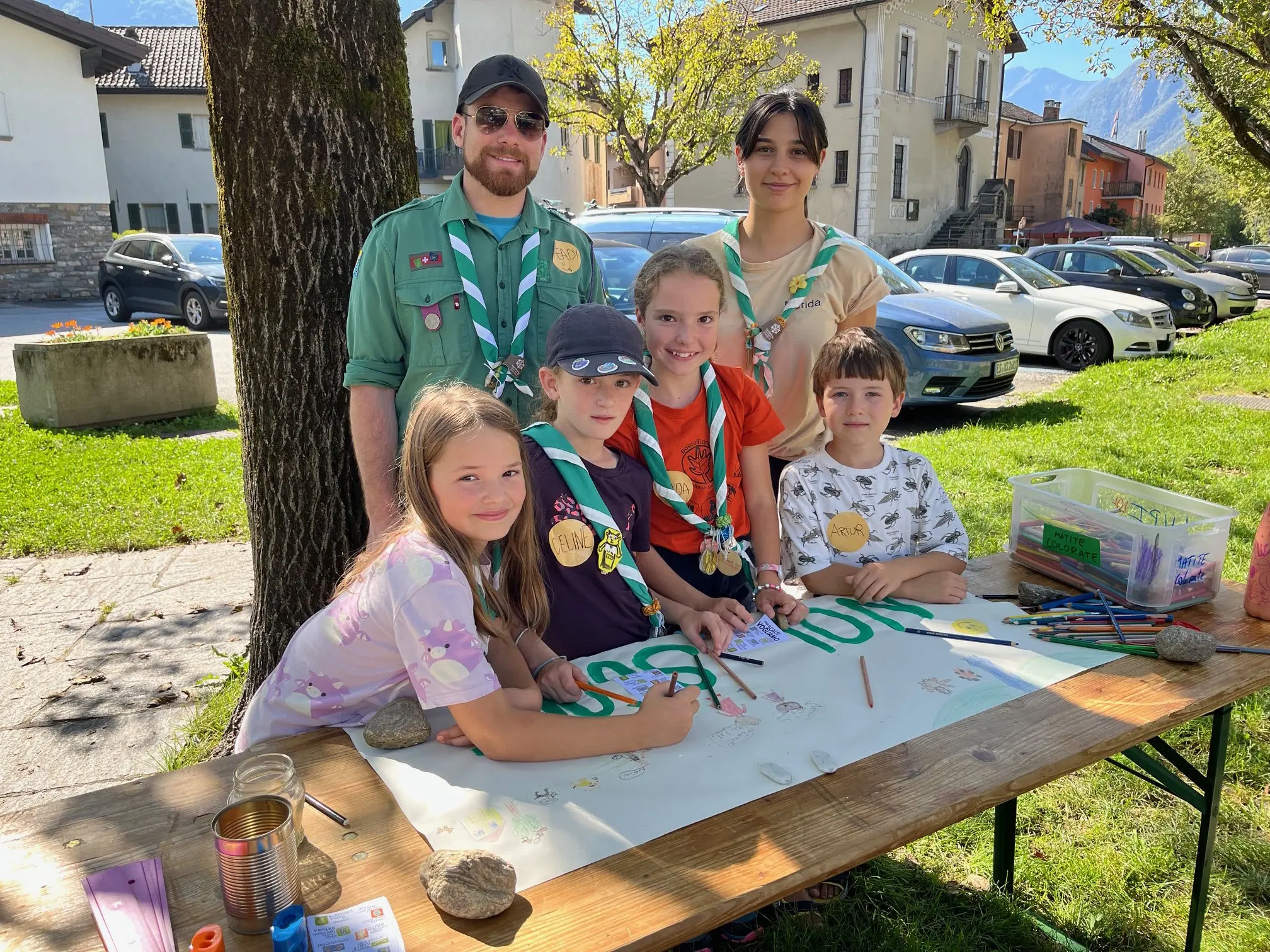Gruppo di ragazzi e adulti sorridenti, coinvolti in un'attività creativa all'aperto. Un tavolo è coperto di materiali per il disegno e cartelloni decorati. Sullo sfondo, case e alberi in un ambiente sereno.