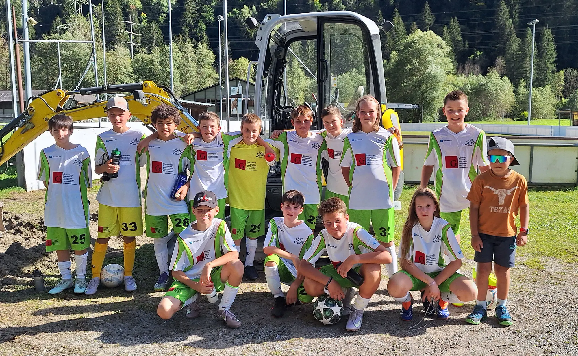 Un gruppo di ragazzi in divisa da calcio sorride davanti a un escavatore. Sono dodici, con palloni da calcio e abbigliamento sportivo. Sullo sfondo ci sono alberi e strutture sportive.