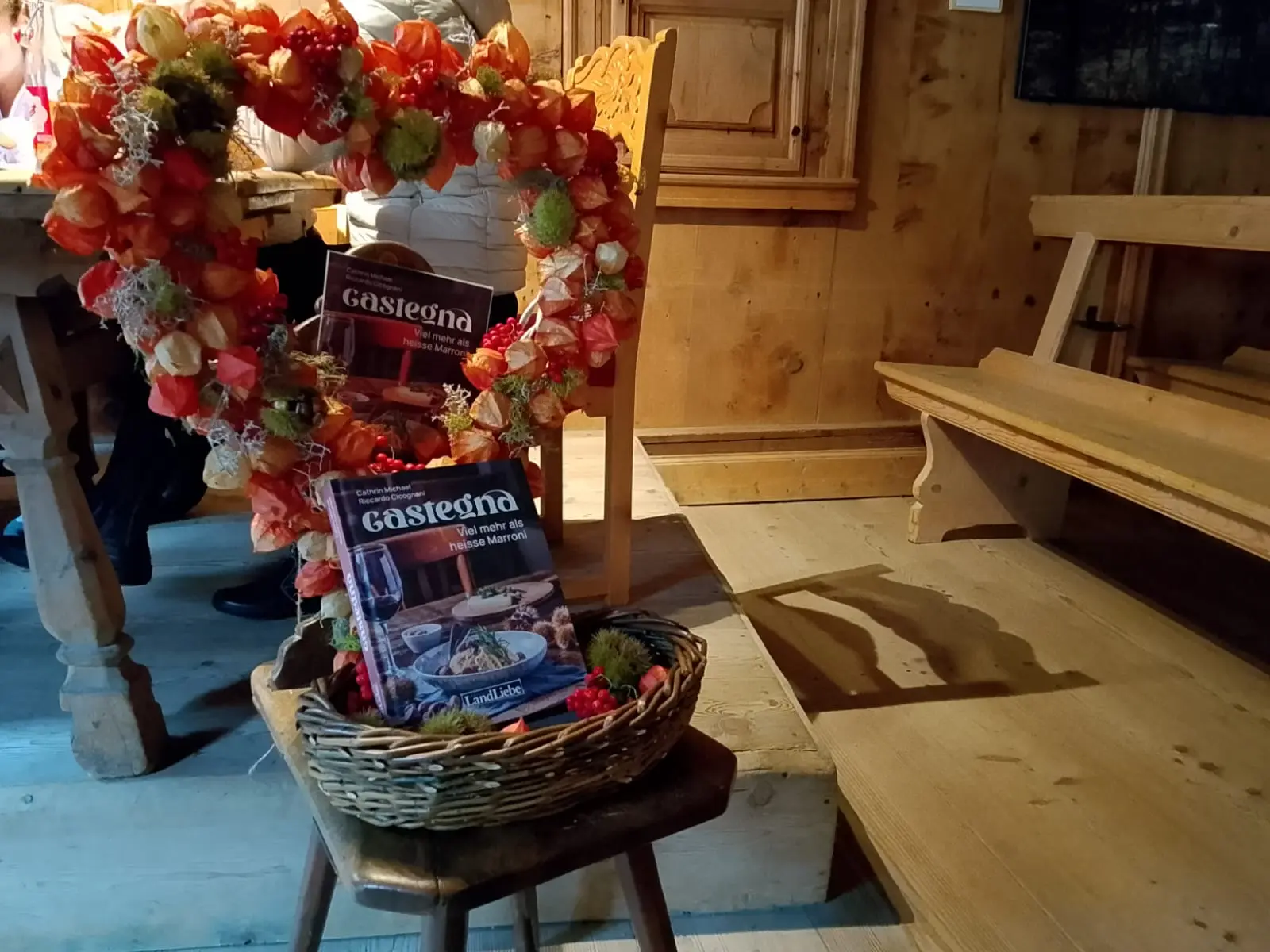 Un cesto di vimini con un libro di cucina dedicato alla castagna; decorato con un guirlanda di fiori colorati. Sullo sfondo si vede un tavolo in legno e un ambiente rustico.