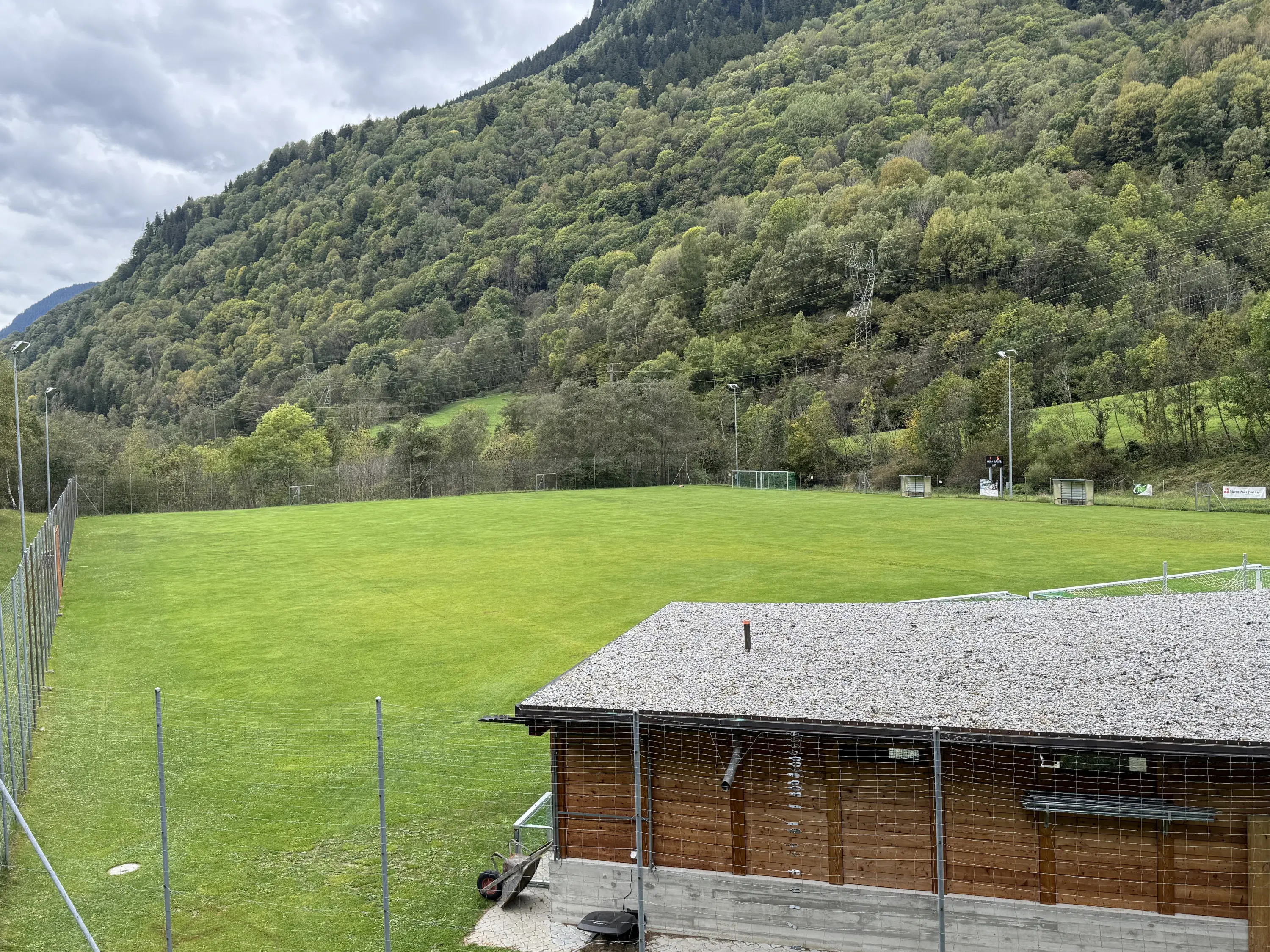 Campo da calcio circondato da colline verdi e alberi. In primo piano, una struttura in legno con un tetto di tegole grigie. Il cielo è nuvoloso.