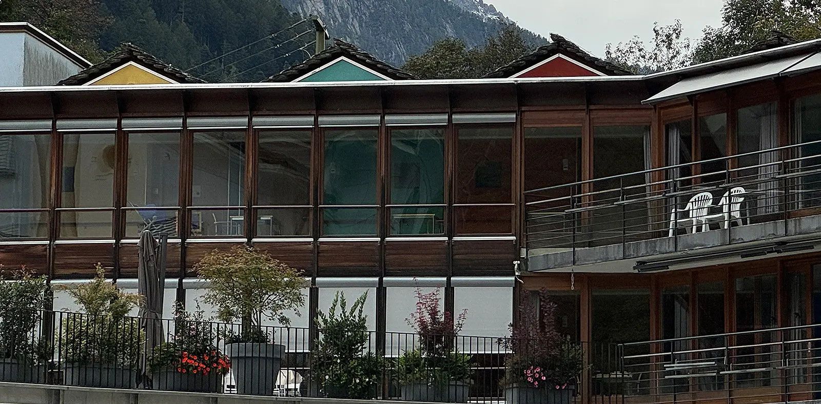 Edificio moderno con facciata in vetro e legno, caratterizzato da tetti colorati e terrazze con piante. Montagne sullo sfondo.