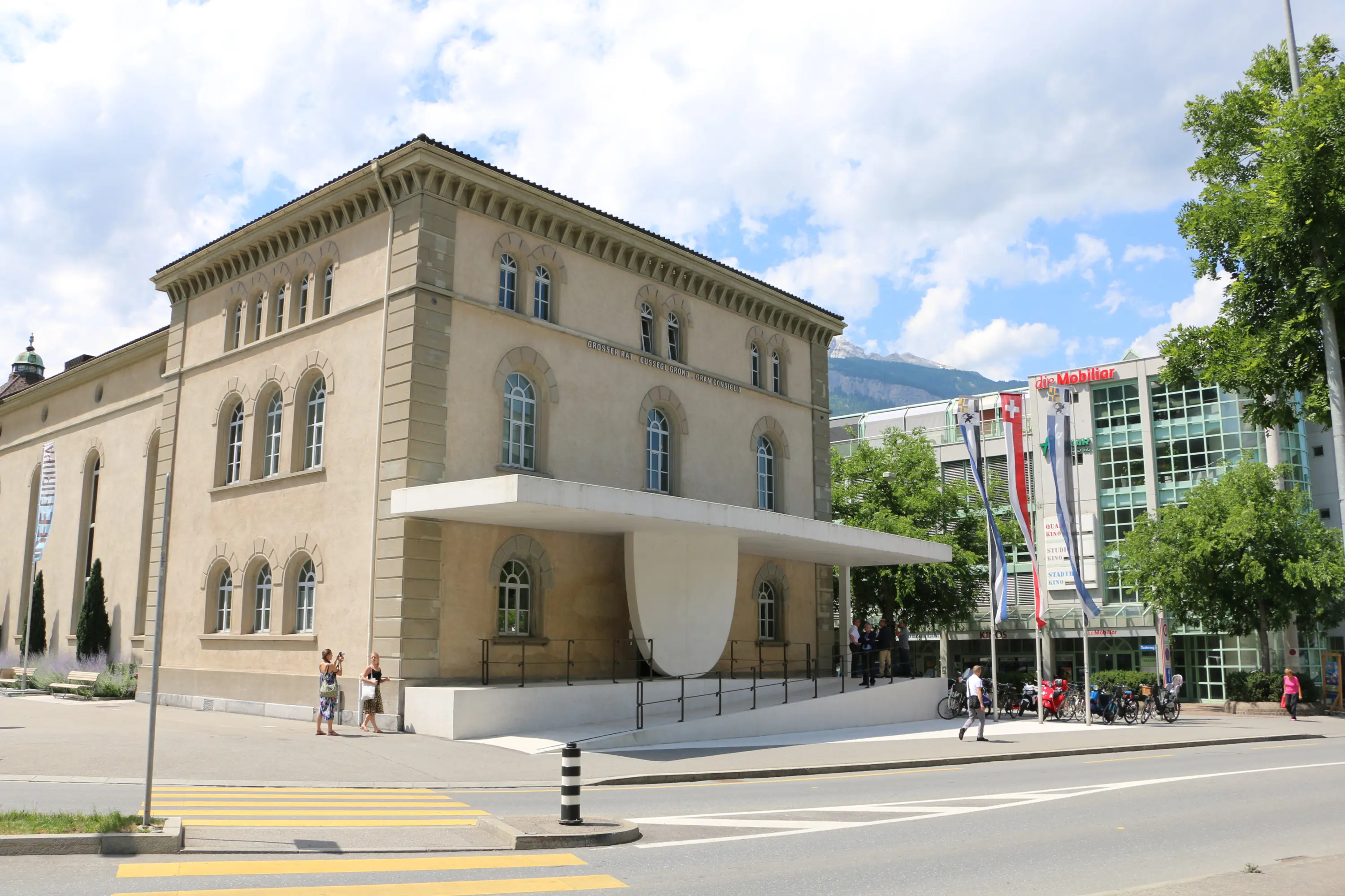 Un edificio storico con facciata in pietra e un grande portico bianco. Sullo sfondo si vedono alberi e altri edifici moderni. Persone passeggiano lungo la strada.