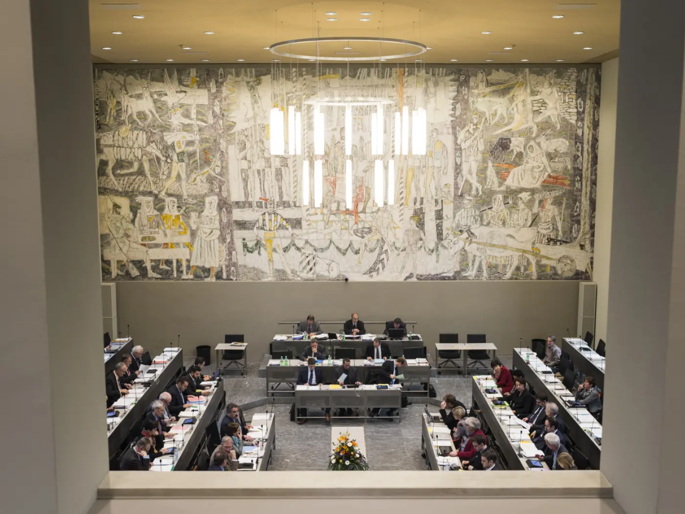 La sala di un'assemblea con persone sedute a tavoli, mentre discutono. Sullo sfondo, un grande murale artistico con figure e motivi complessi. Una lampada a sospensione illumina l'ambiente.