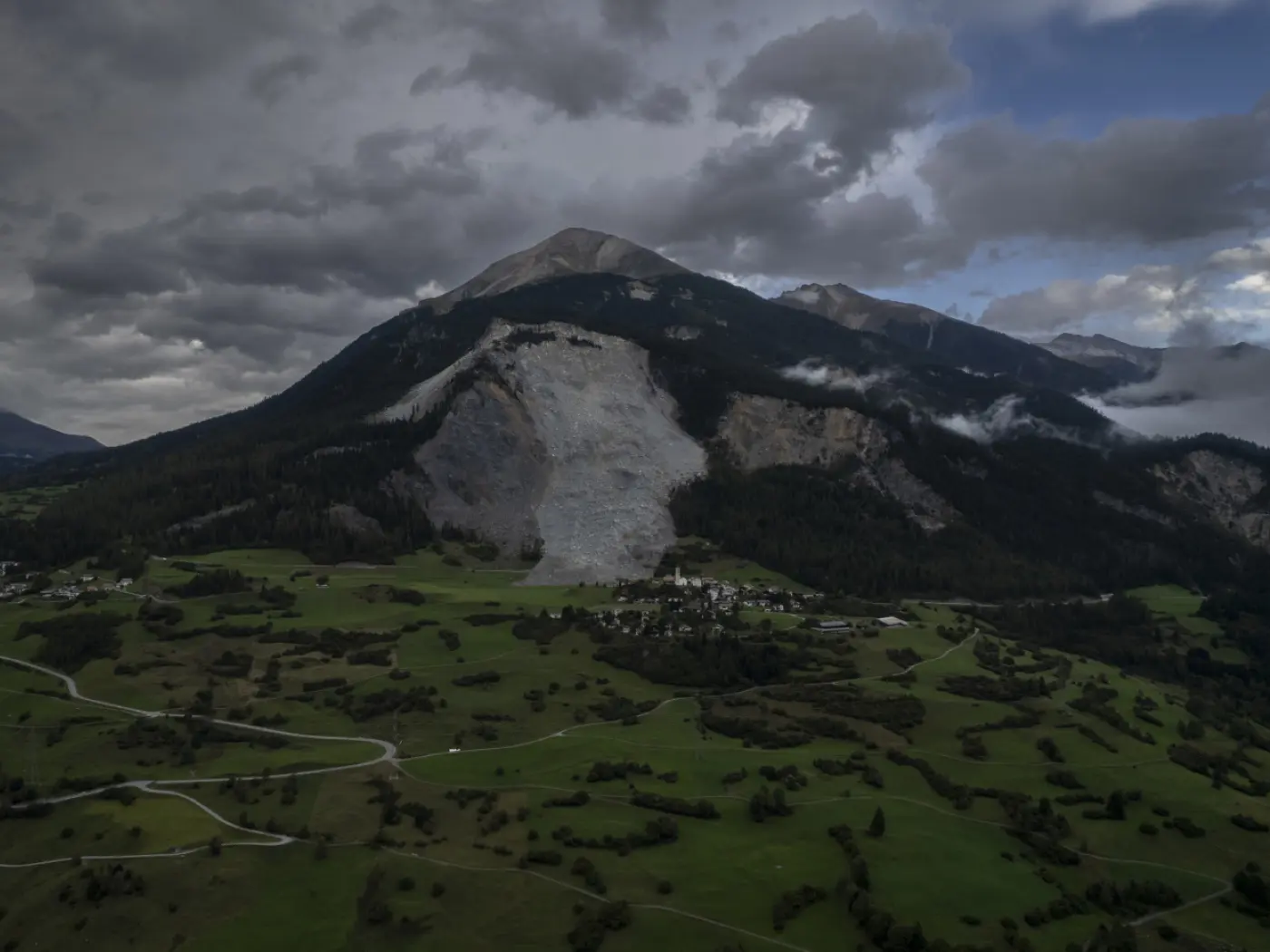 Un'imponente frana si è staccata da una montagna, lasciando una vasta area rocciosa esposta. Sullo sfondo, nuvole scure si addensano sopra il paesaggio verdeggiante e collinare sottostante.
