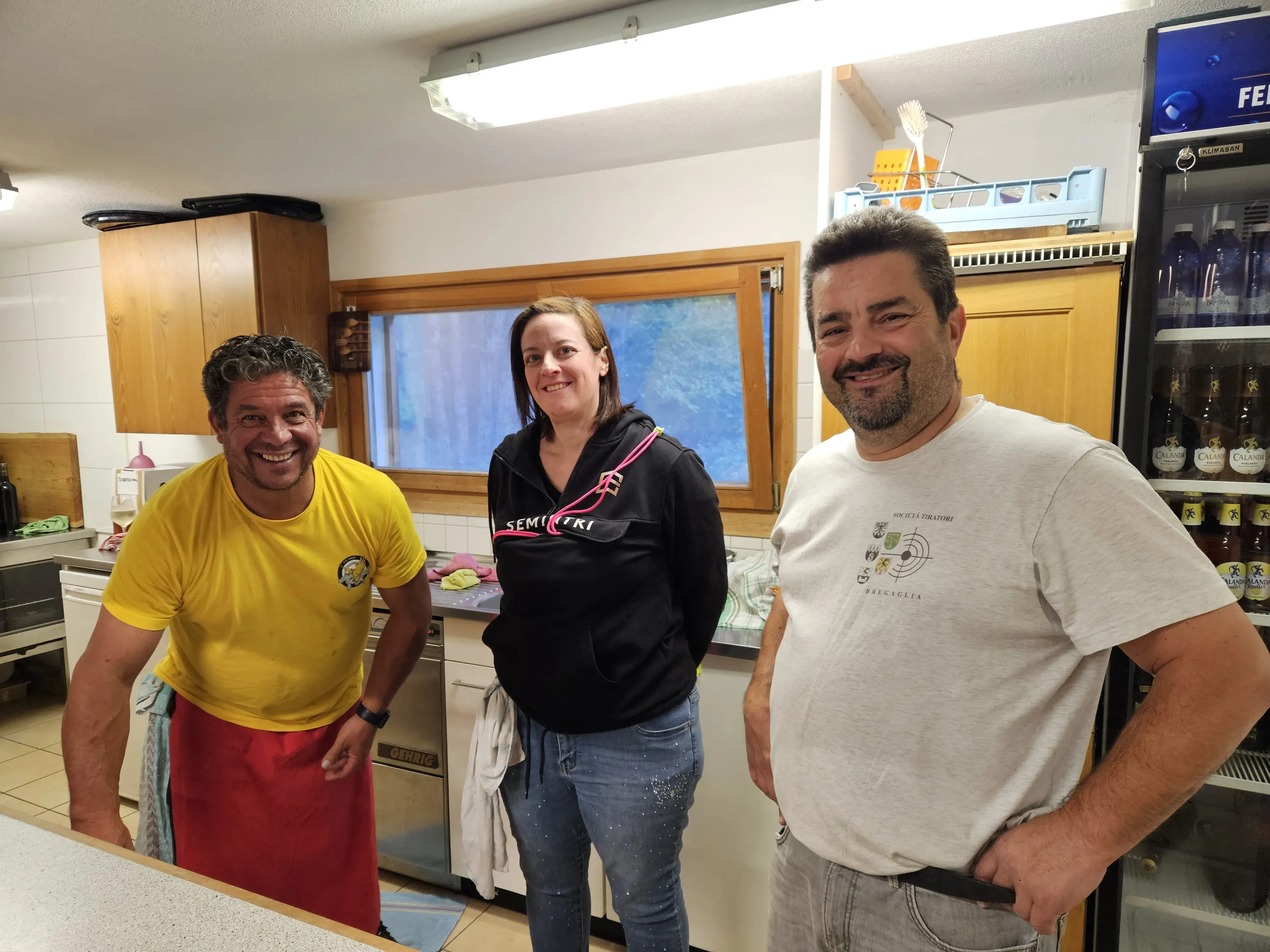 Tre persone sorridenti in una cucina. Un uomo con una maglietta gialla e un grembiule rosso, una donna con una felpa nera e un uomo con una maglietta chiara. Tutti e tre sono in ottima disposizione, circondati da attrezzature da cucina.