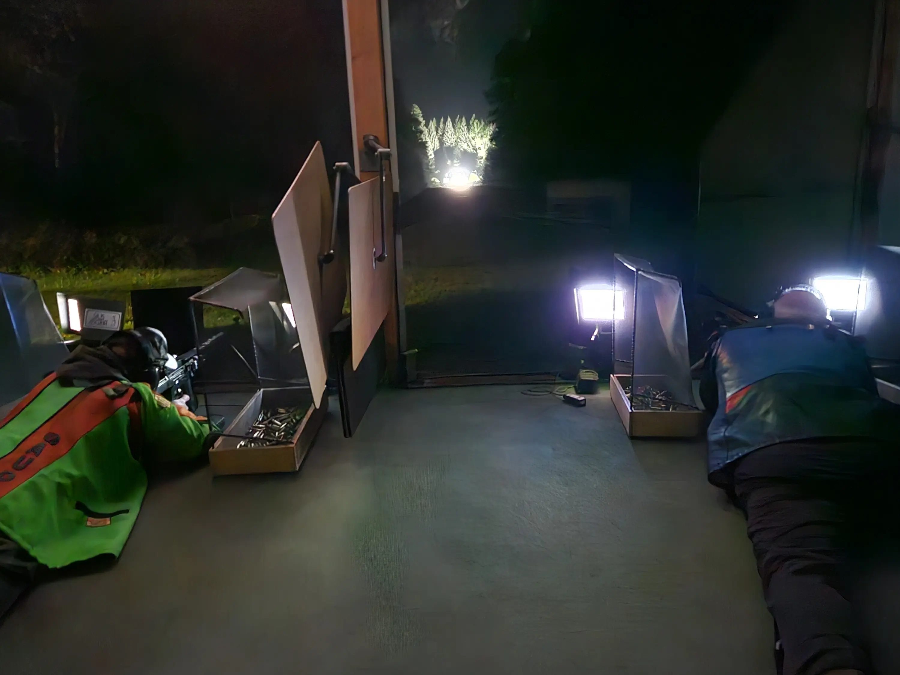 Due uomini si trovano chinati in un ambiente scuro, intenti a lavorare con strumenti luminosi. Alle loro spalle ci sono scatole aperte e luci artificiali che illuminano il terreno. In lontananza, si intravede una fonte di luce proveniente da un'area boscosa.