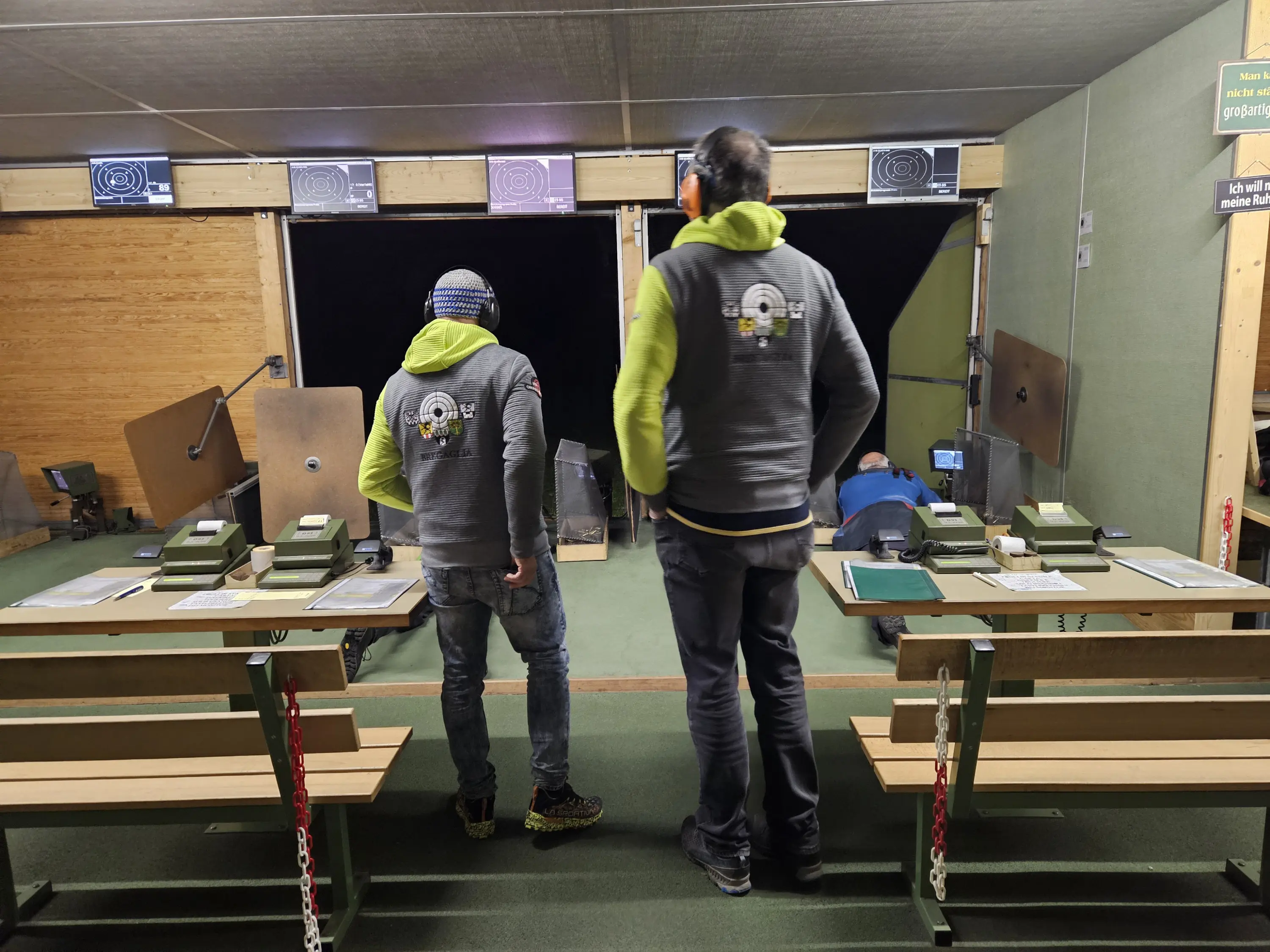 Due uomini in maglietta grigia e cappelli, di spalle, si trovano in una sala di tiro. Davanti a loro ci sono postazioni di tiro con bersagli, mentre un terzo uomo è chinato a terra. Banco e attrezzature sono visibili sul lato.