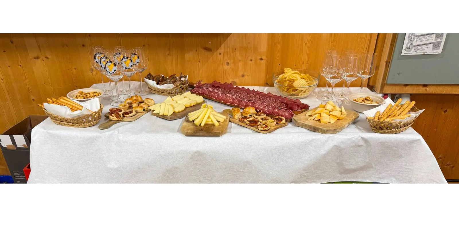 Tavolo imbandito con un assortimento di salumi, formaggi, snack e dolci, accompagnato da calici di vetro.