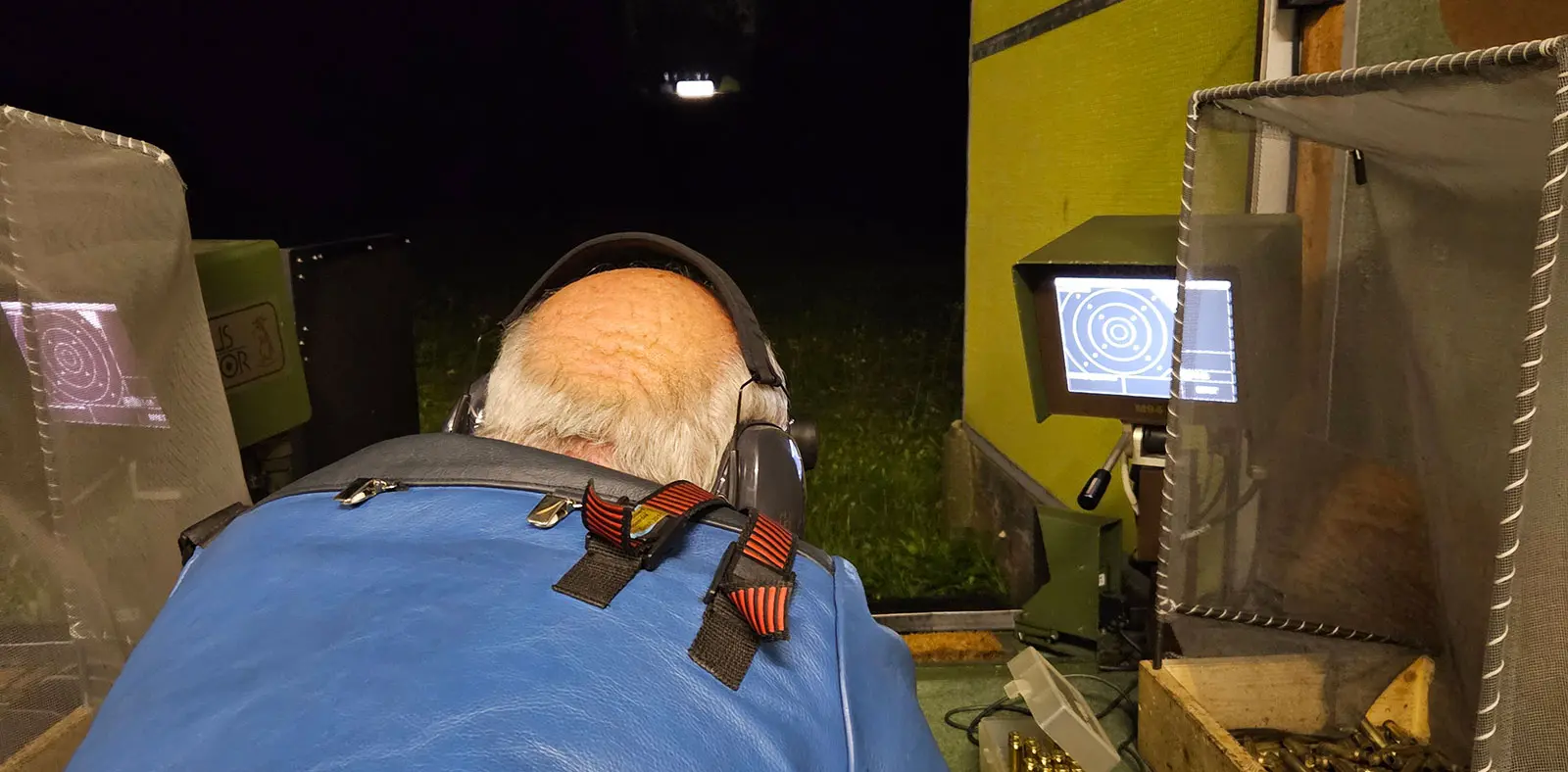 Un uomo con una giacca azzurra è seduto di spalle, mentre utilizza un sistema di mira elettronico. Il campo è buio e ci sono monitor che mostrano dati di target.