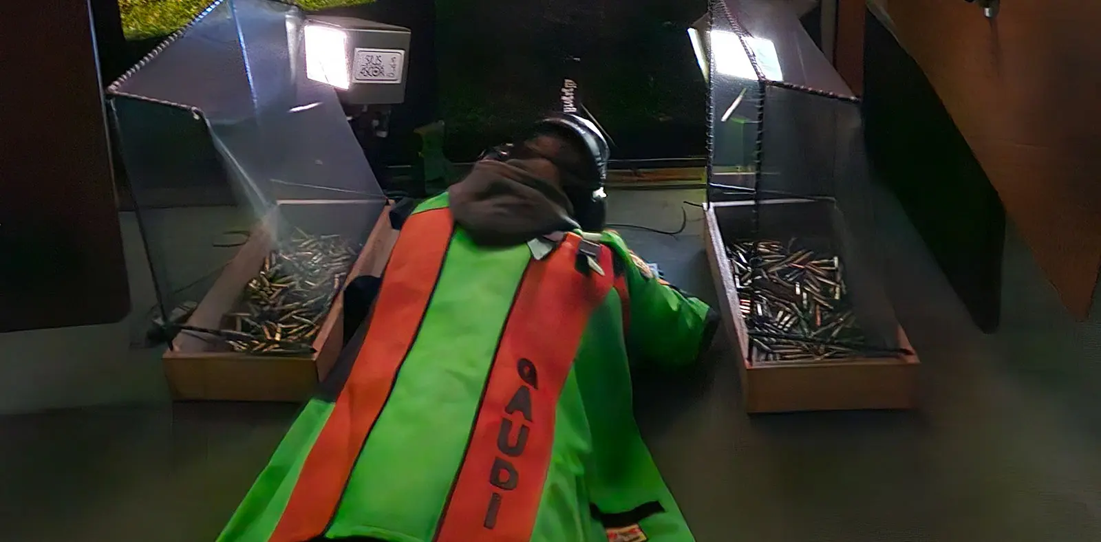 Un uomo disteso su un pavimento, indossando un giubbotto ad alta visibilità verde e arancione, circondato da scatole trasparenti contenenti proiettili. Luce proveniente da dietro illumina parzialmente la scena.