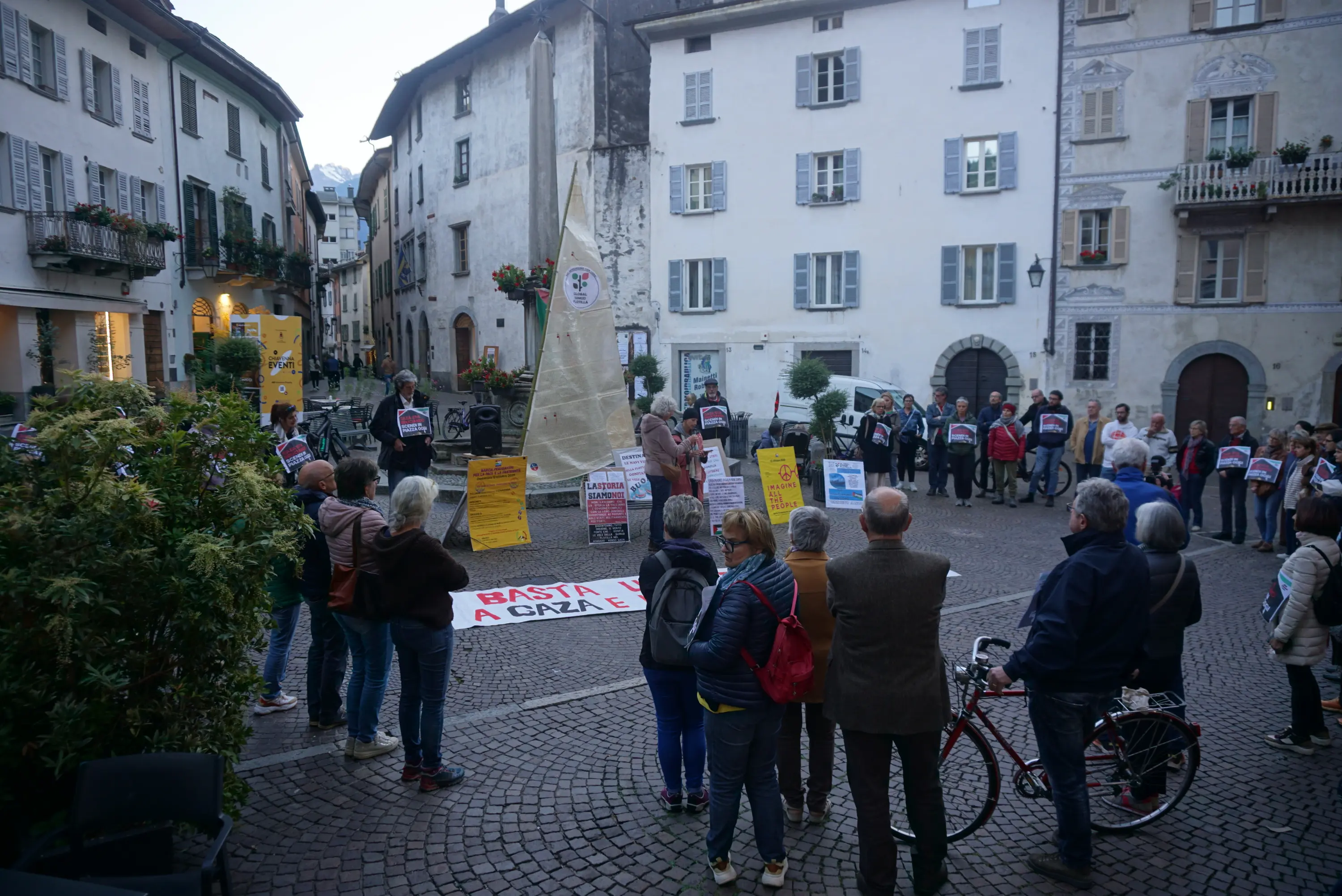 Un gruppo di persone si raduna in una piazza con cartelli e striscioni, manifestando contro un tema locale. Sullo sfondo si vede un edificio storico e alcune biciclette parcheggiate. È sera e l'atmosfera è vivace.