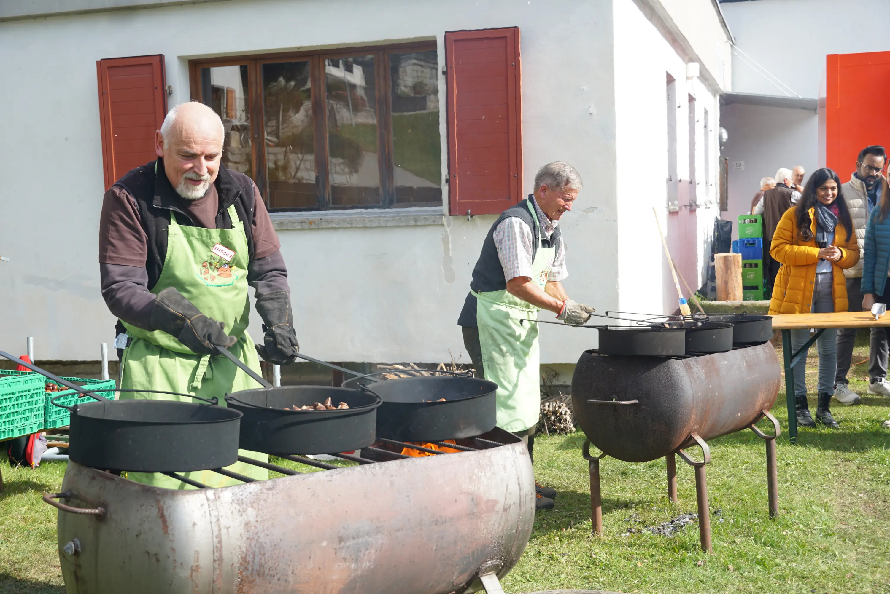 Due uomini, vestiti con grembiuli, cucinano all'aperto su due grandi recipienti di metallo. Si vedono persone in distanza che osservano l'evento. Il sole splende su un ambiente sereno con un edificio bianco sullo sfondo.