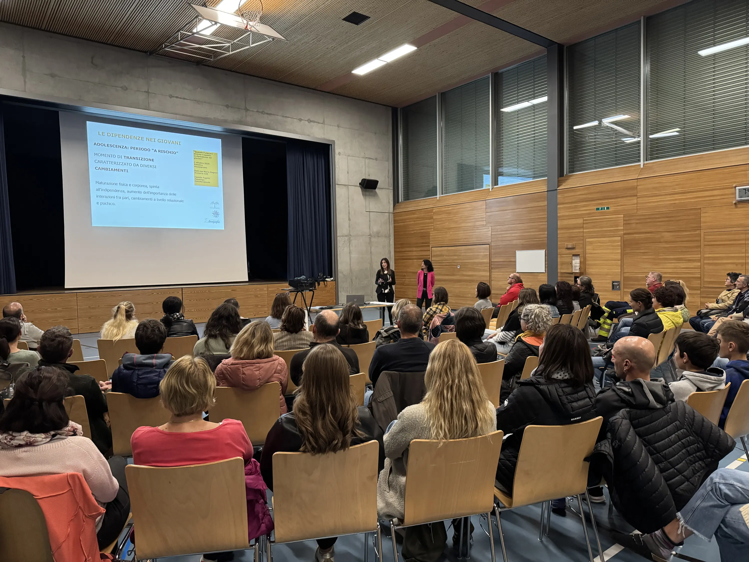 Una presentazione in aula con un pubblico numeroso. Sullo schermo è visibile una diapositiva con del testo. Le persone sono sedute su sedie in legno, attentamente rivolte verso il relatore.