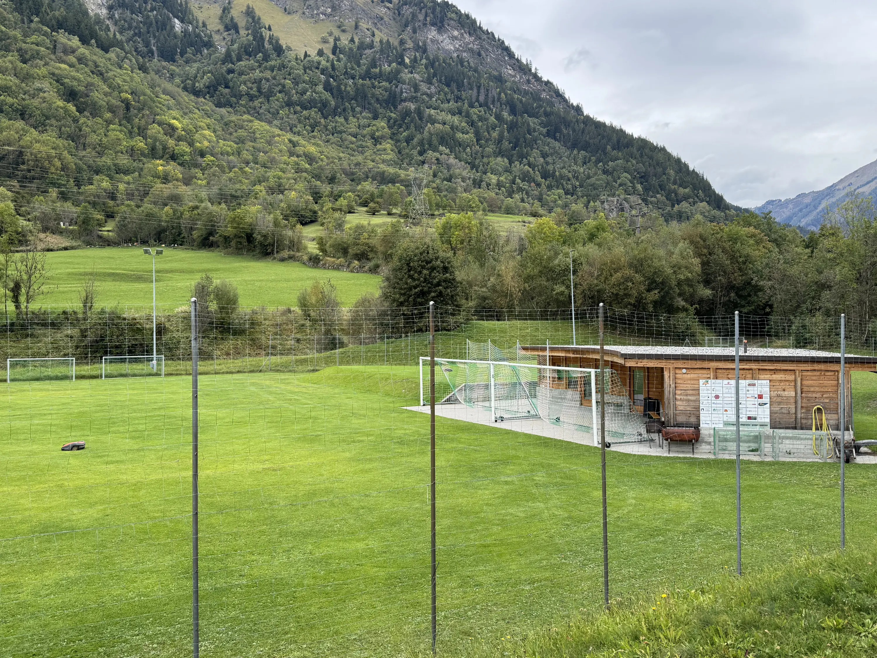 Un campo sportivo di calcio circondato da colline verdi, con un edificio in legno adiacente. In lontananza si vedono alberi e montagne. Il cielo è nuvoloso.