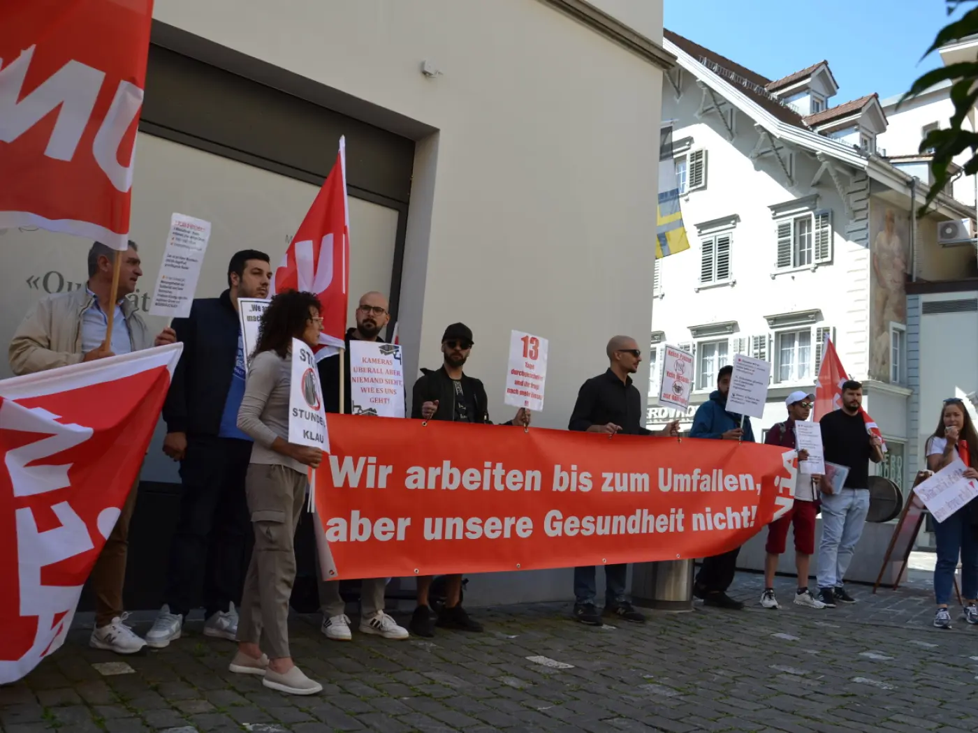 Manifestazione con persone che tengono cartelli e striscioni, protestando per i diritti dei lavoratori. I partecipanti indossano t-shirt e mostrano bandiere rosse. Un grande striscione recita "Wir arbeiten bis zum Umfallen, aber unsere Gesundheit nicht!"