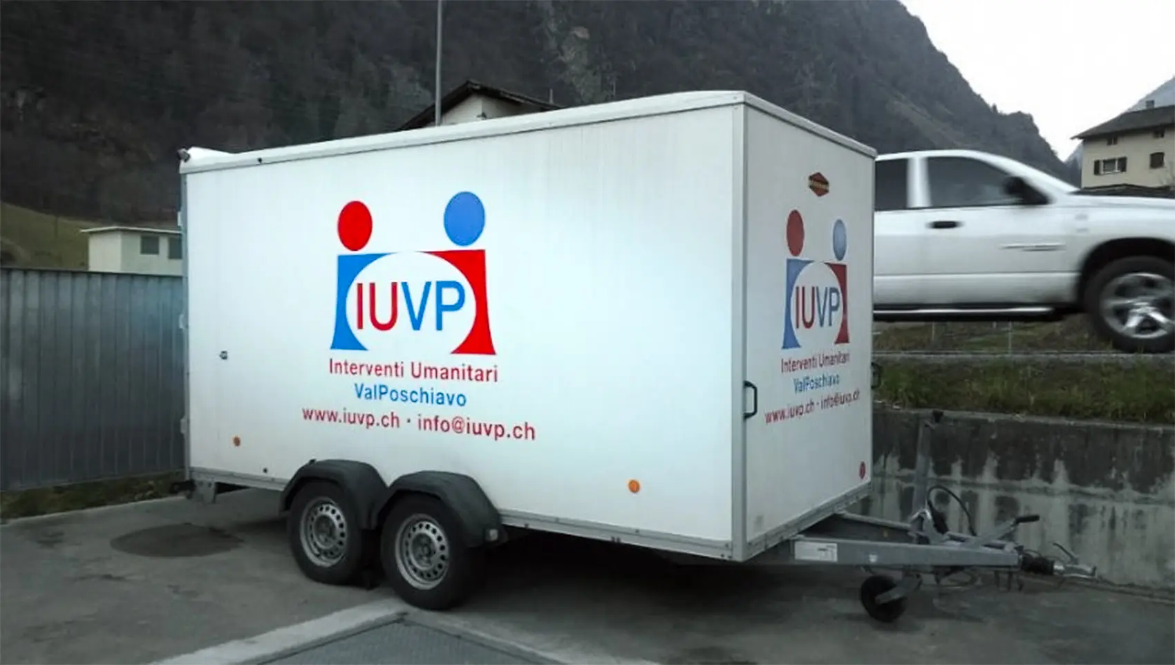 Un rimorchio bianco con il logo "IUVP" e la scritta "Interventi Umanitari ValPoschiavo" su un lato, parcheggiato in un'area all'aperto con montagne sullo sfondo.