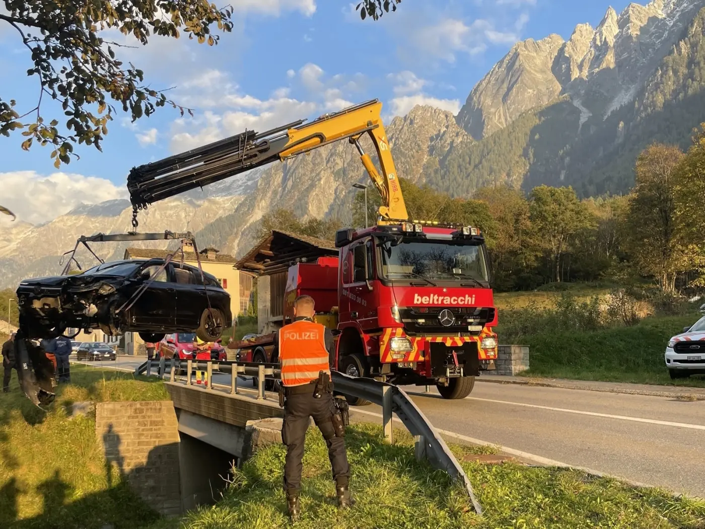Un camion della società Beltracchi solleva un'auto nera avvolta in una corda, mentre un uomo in divisa della polizia osserva. Sullo sfondo si vedono montagne e un cielo azzurro.