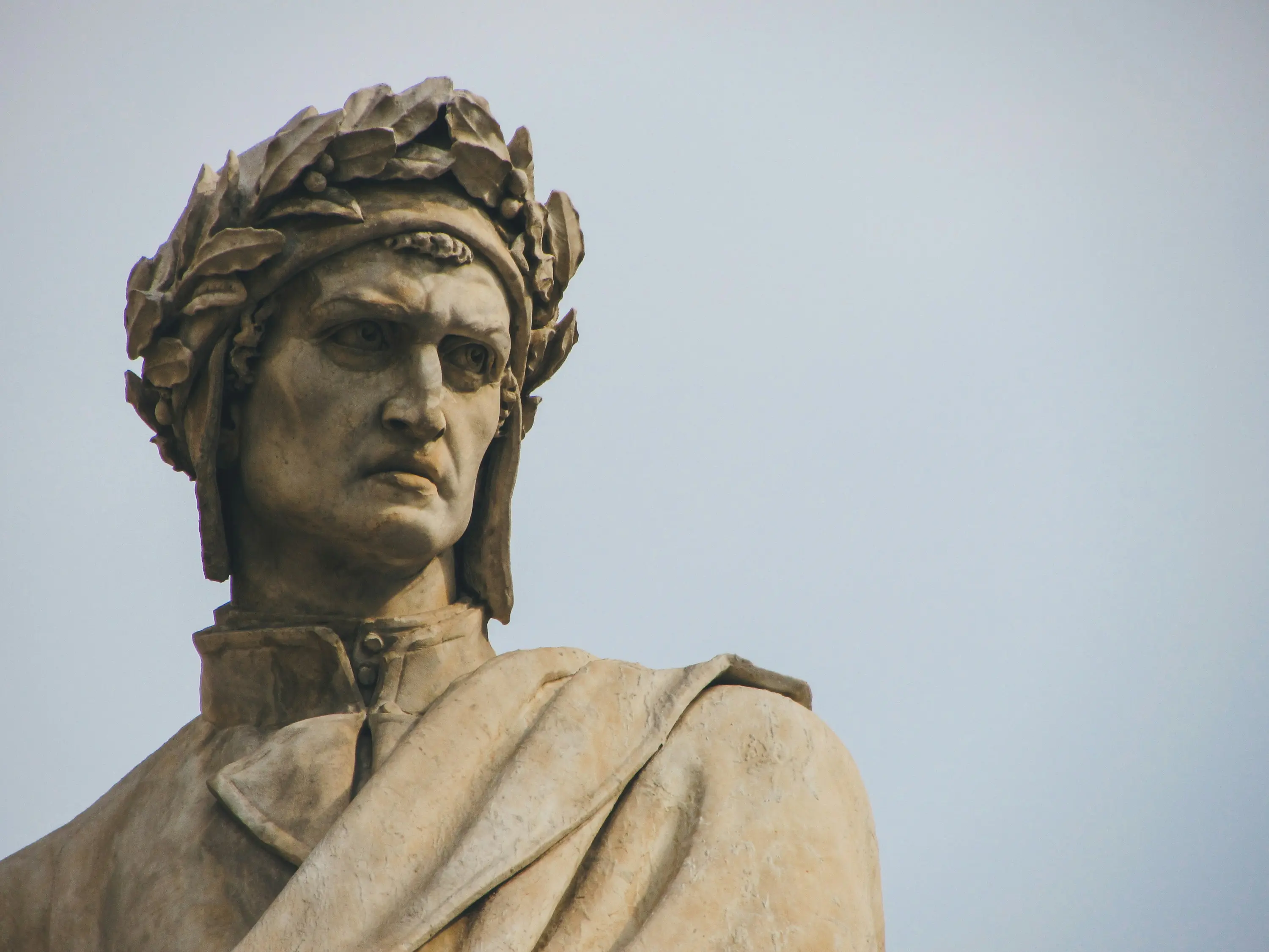 Statua di Dante Alighieri con corona di alloro, caratterizzata da un'espressione seria e dettagliata, posizionata su uno sfondo chiaro.